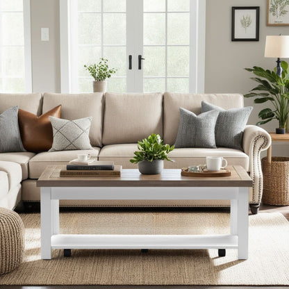 A cozy living room with a beige sofa, gray and brown pillows, and the Hampton Coffee Table in Jasmine Whitewash/Barnwood, topped with books, a plant, and mugs on a woven rug by large windows streaming natural light.