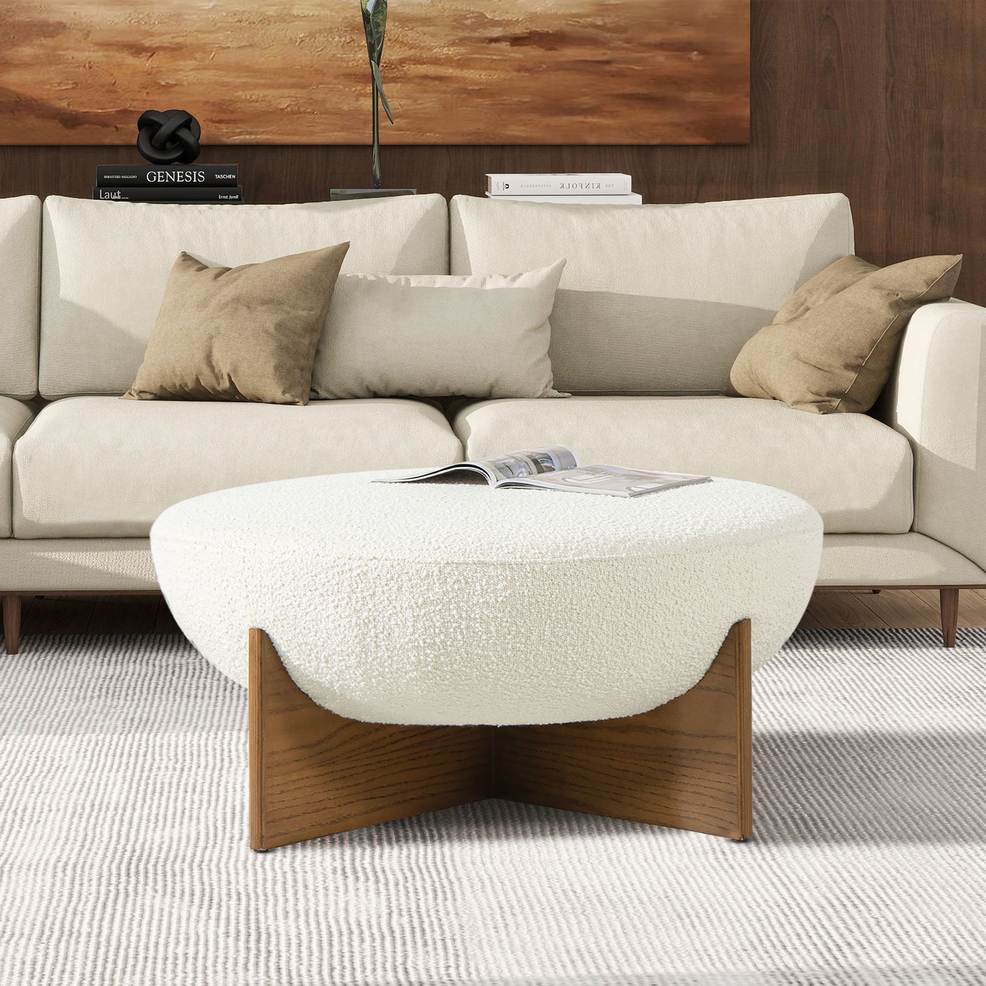 A modern living room featuring a cream sofa, brown and beige pillows, the Bali Round Upholstered Ottoman with a wooden base and an open book on top, plus a neutral rug on the floor.