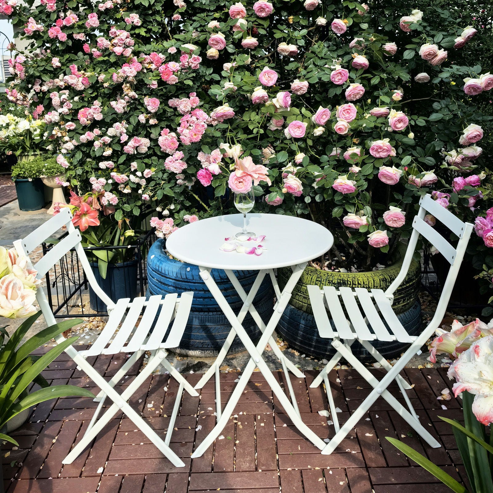 The 3 Piece Patio Bistro Set of Foldable Round Table and Chairs sits on a brick patio among pink roses, with a glass and a vase of roses on the table, creating a peaceful garden scene.