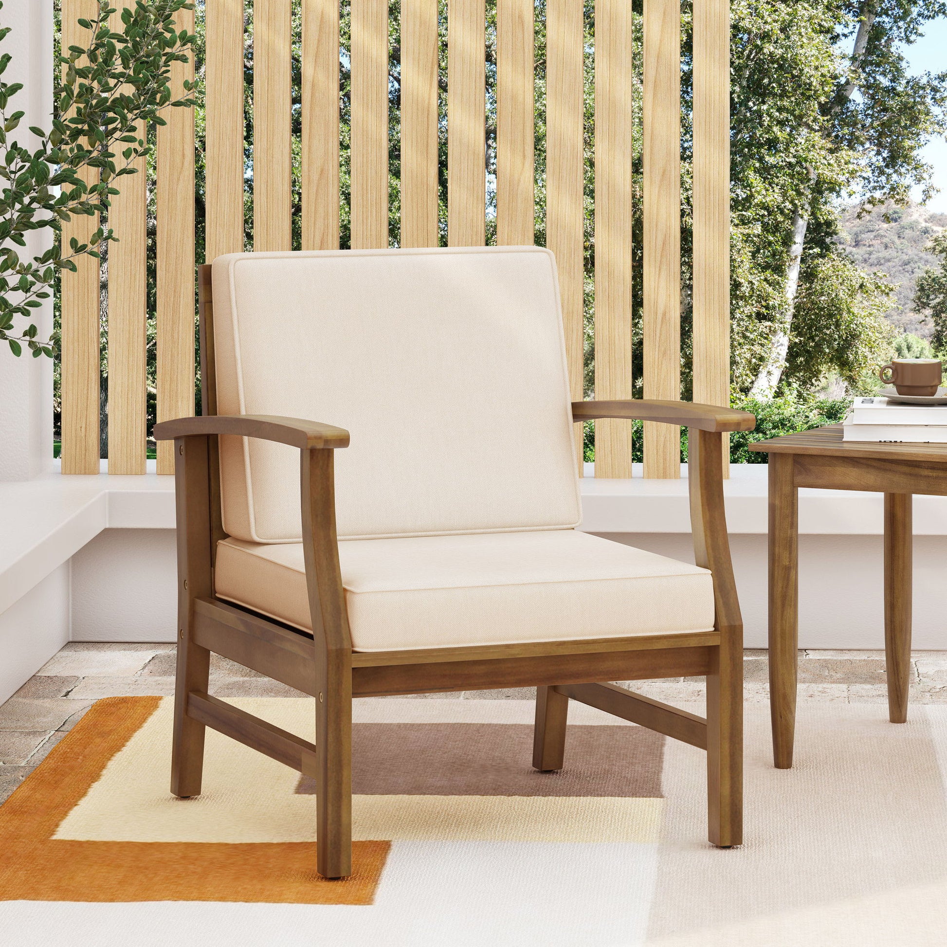The Perla Contemporary Club Chair with water resistant cushions sits on a patterned rug outdoors beside a wooden table, framed by vertical wooden slats and greenery in the background.