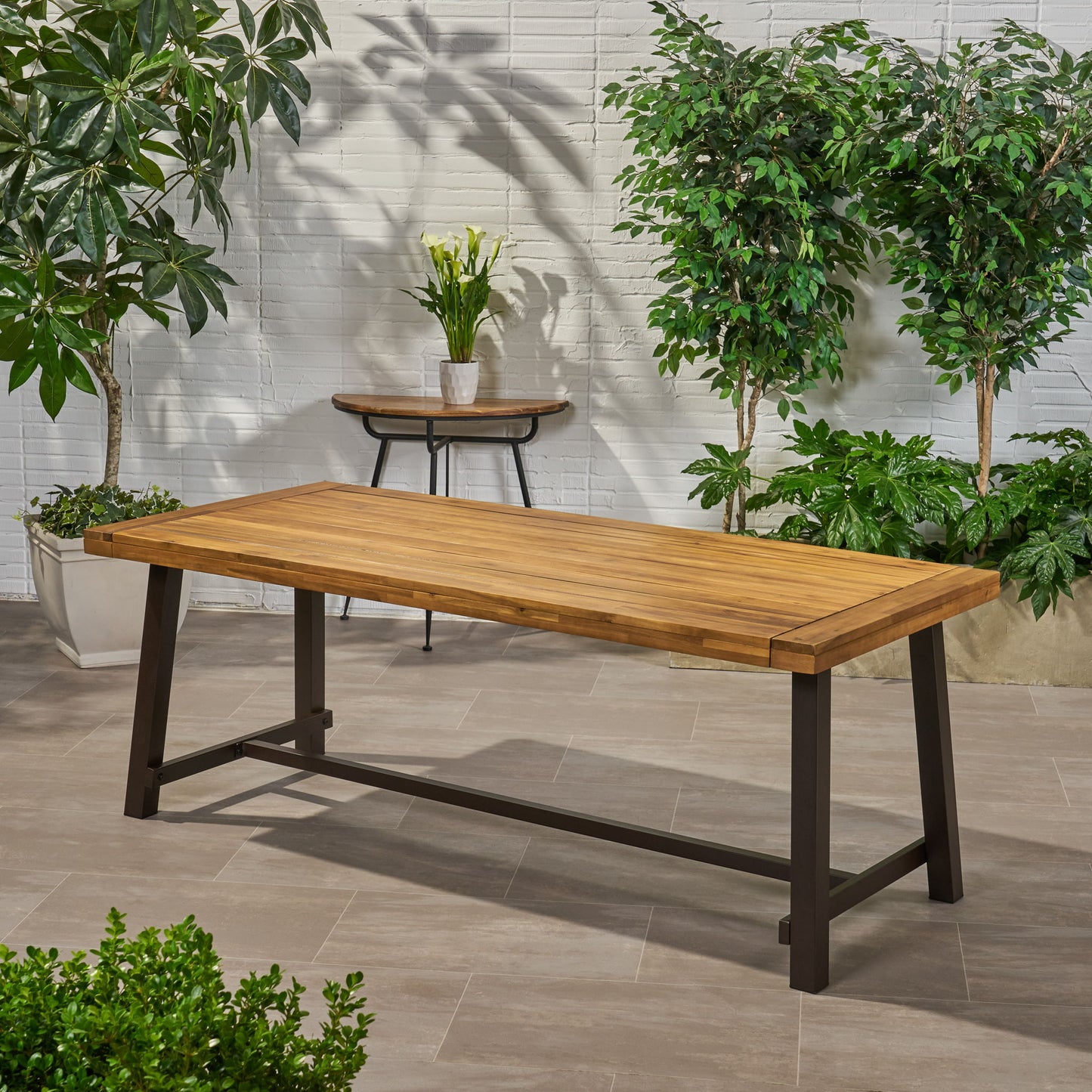 The Equilibrium Outdoor Dining Table with a wooden frame and slat top stands on a tiled patio, surrounded by green potted plants and a side table with a white vase of flowers against a white brick wall.