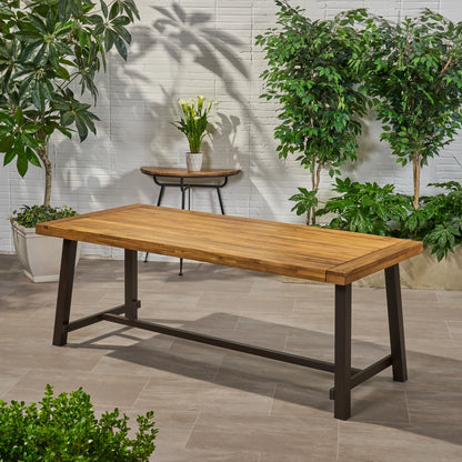 The Equilibrium Outdoor Dining Table with a wooden frame and slat top stands on a tiled patio, surrounded by green potted plants and a side table with a white vase of flowers against a white brick wall.