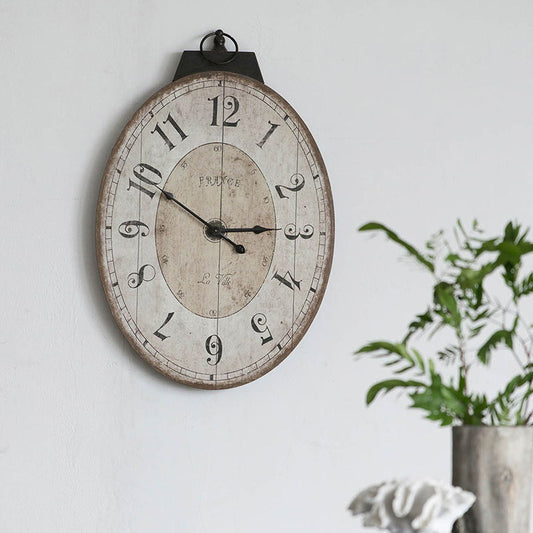 An Antique Oval Wall Clock, Traditional Vintage Home Decor Clock - Ivory hangs on a white wall, featuring large black numbers and a distressed white face. Green leafy plants in vases appear on the right side of the scene.