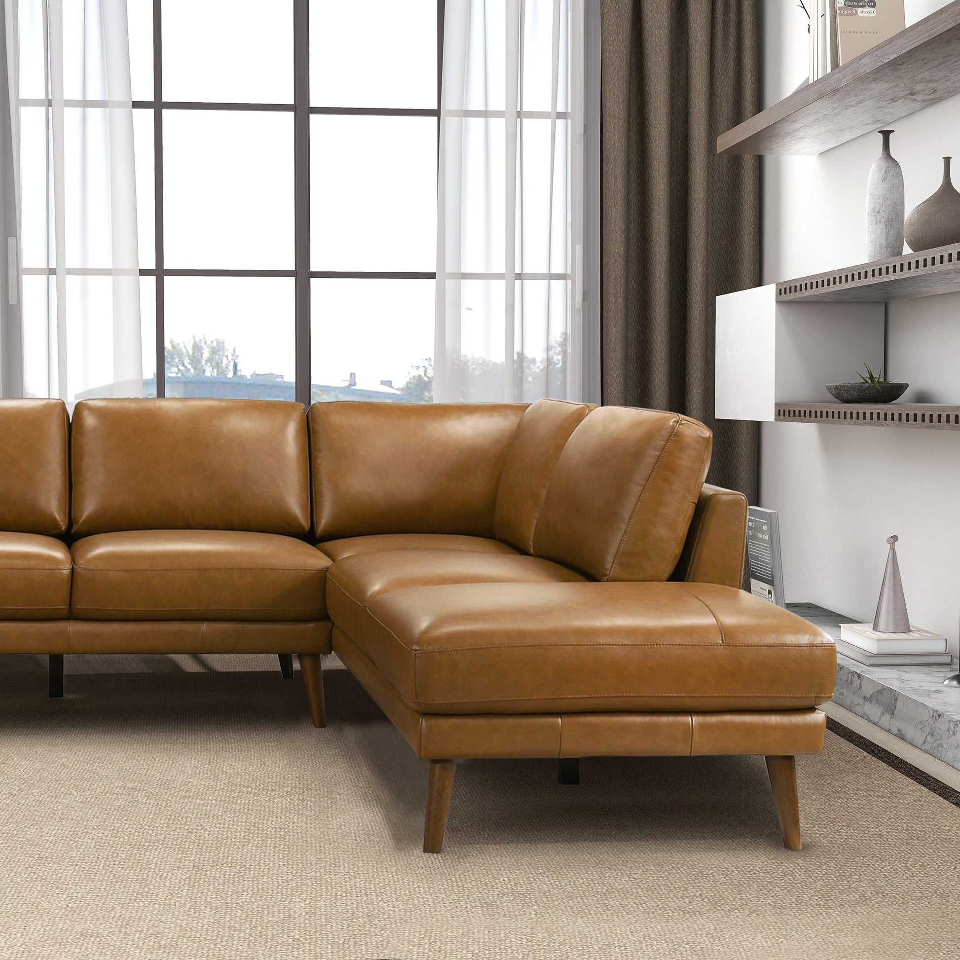 A modern living room featuring the Benson - Sectional Sofa With Chaise in brown leather on a beige carpet, with a large curtained window behind and white shelves with decorative items on the right.
