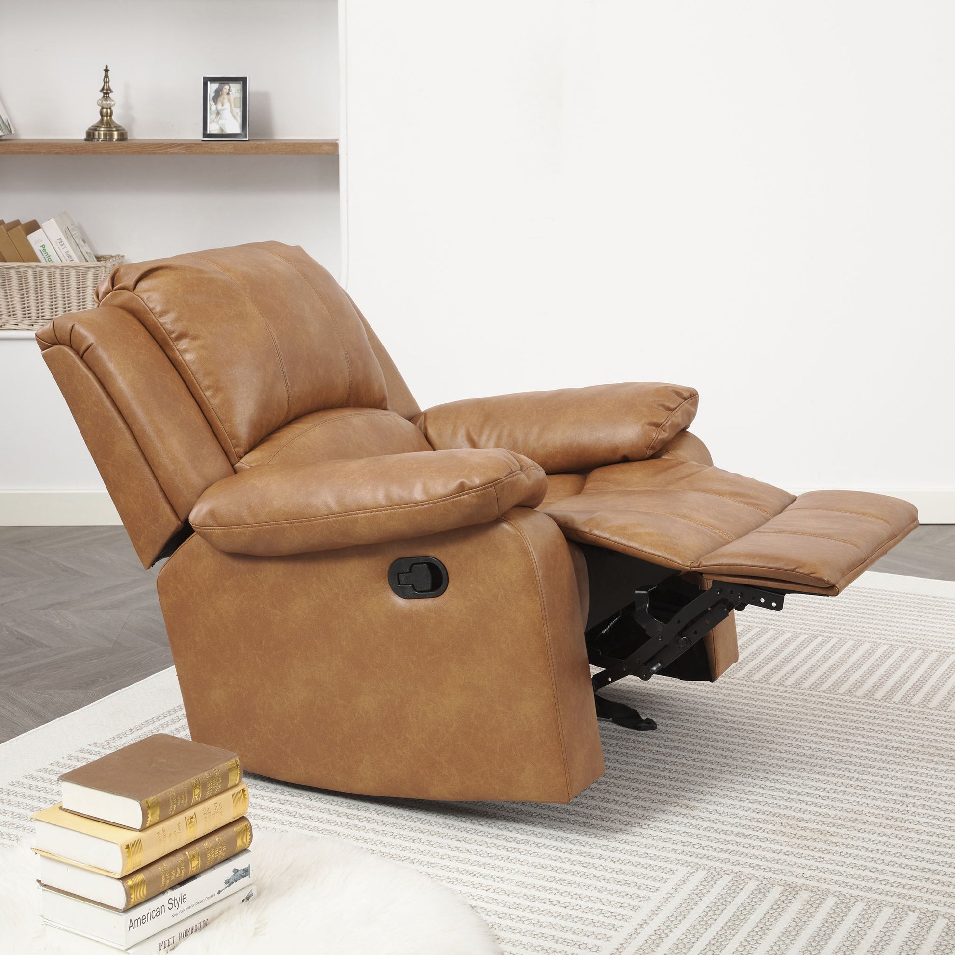 The Charlotte Glider Recliner With Classic Style in tan leather, featuring an extended footrest, sits on a light patterned rug in a modern living room with books and decor displayed on shelving behind it.