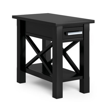 The Kitchener Handcrafted Table is a black wooden side table featuring a single drawer with a silver handle, crossed side supports, and a lower shelf, shown against a plain white background.