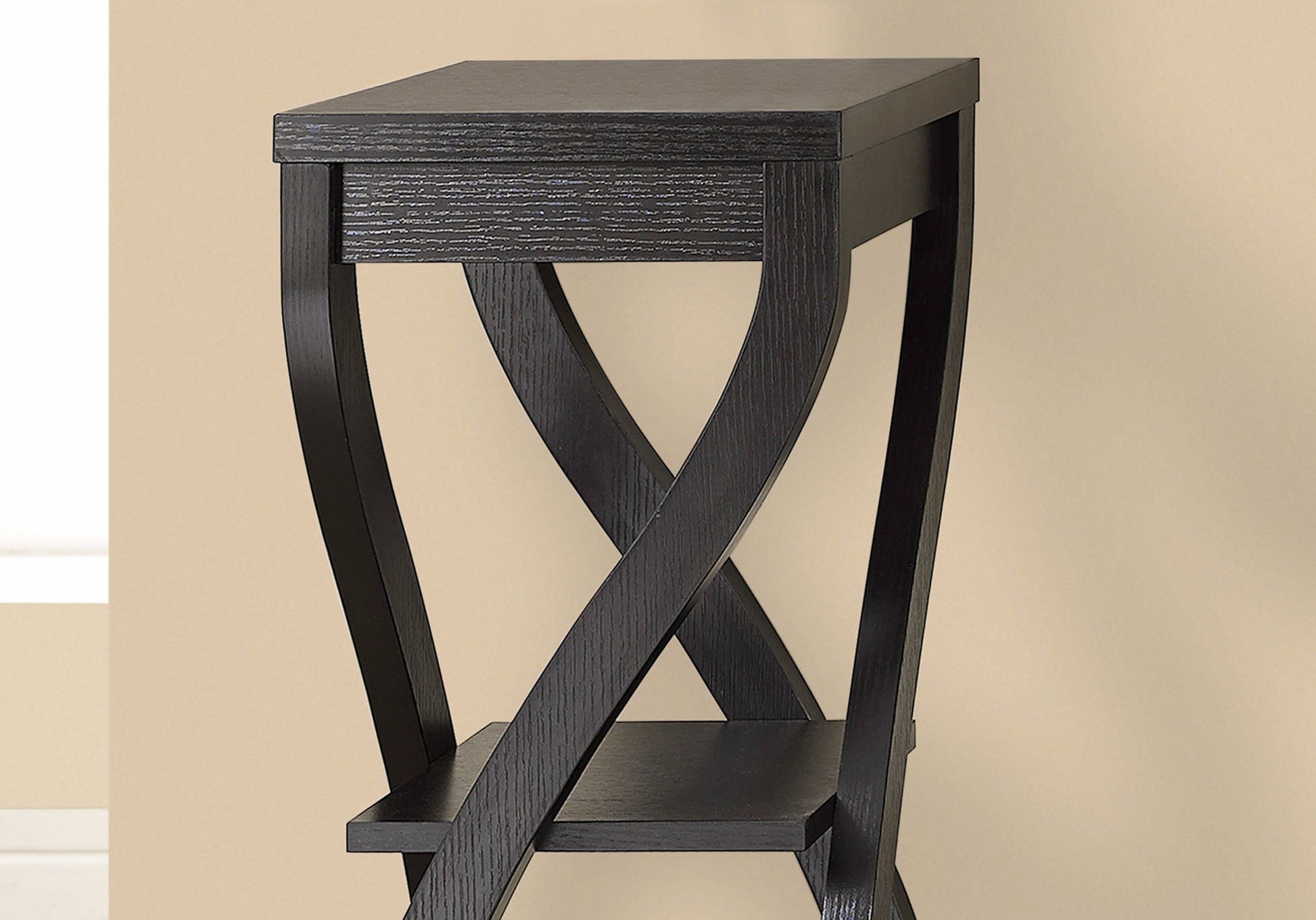 A contemporary Accent Table with a square top and X-shaped legs, featuring versatile usage and a lower shelf, is placed against a beige wall.
