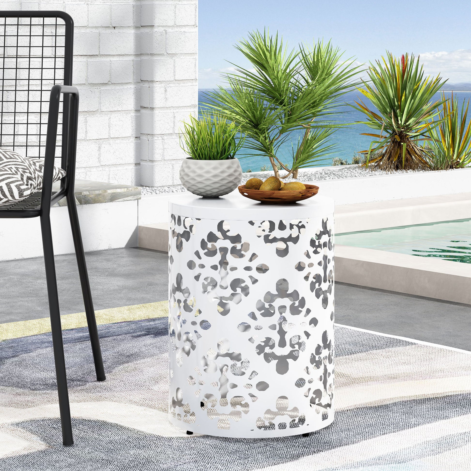 The Outdoor Metal Modern Design End Table, featuring a cut-out pattern, holds a potted plant and a bowl of snacks by the poolside patio, with tropical plants and a coastal view in the background.
