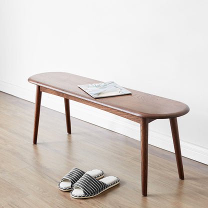 The Dining Bench, Table Bench For Living Room with rounded edges stands against a white wall on a hardwood floor. A magazine lies on the bench, and striped slippers are placed on the floor in front of it.