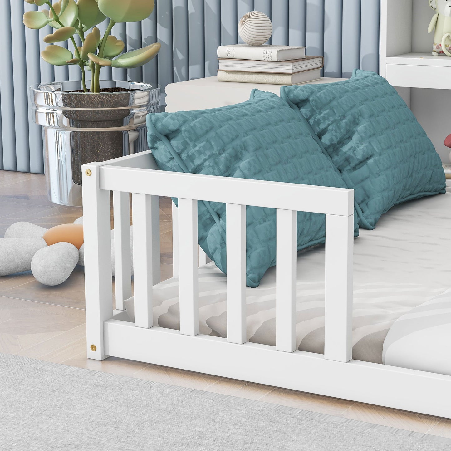 A Floor Bed With Side Bookcase, Shelves, and Guardrails featuring a white wooden frame is paired with two blue textured pillows, near a shiny silver plant pot and soft decorative stones, with vertical blue wall panels behind.