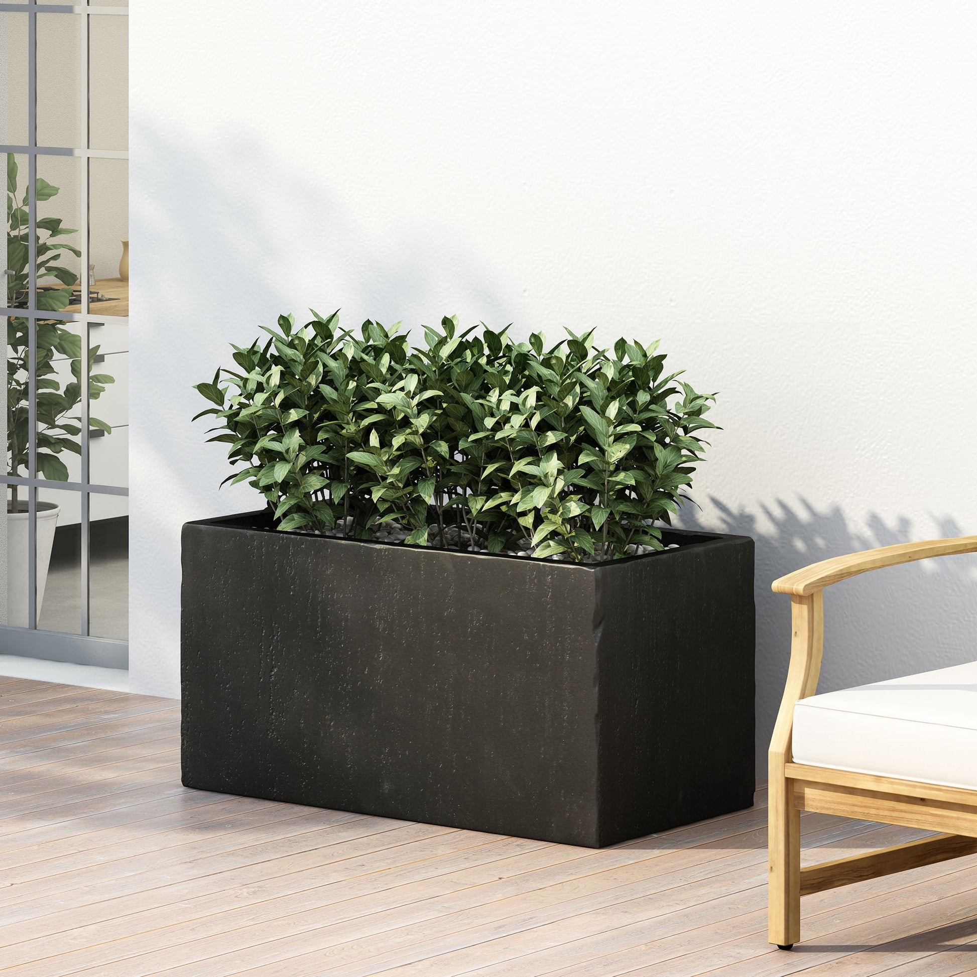 The Modern Outdoor Planter in black, filled with green plants, sits on a wooden deck beside a light wall and an outdoor chair with white cushions. A nearby window reflects indoor greenery.