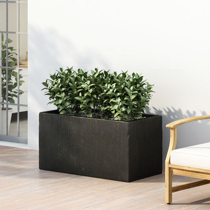 The Modern Outdoor Planter in black, filled with green plants, sits on a wooden deck beside a light wall and an outdoor chair with white cushions. A nearby window reflects indoor greenery.