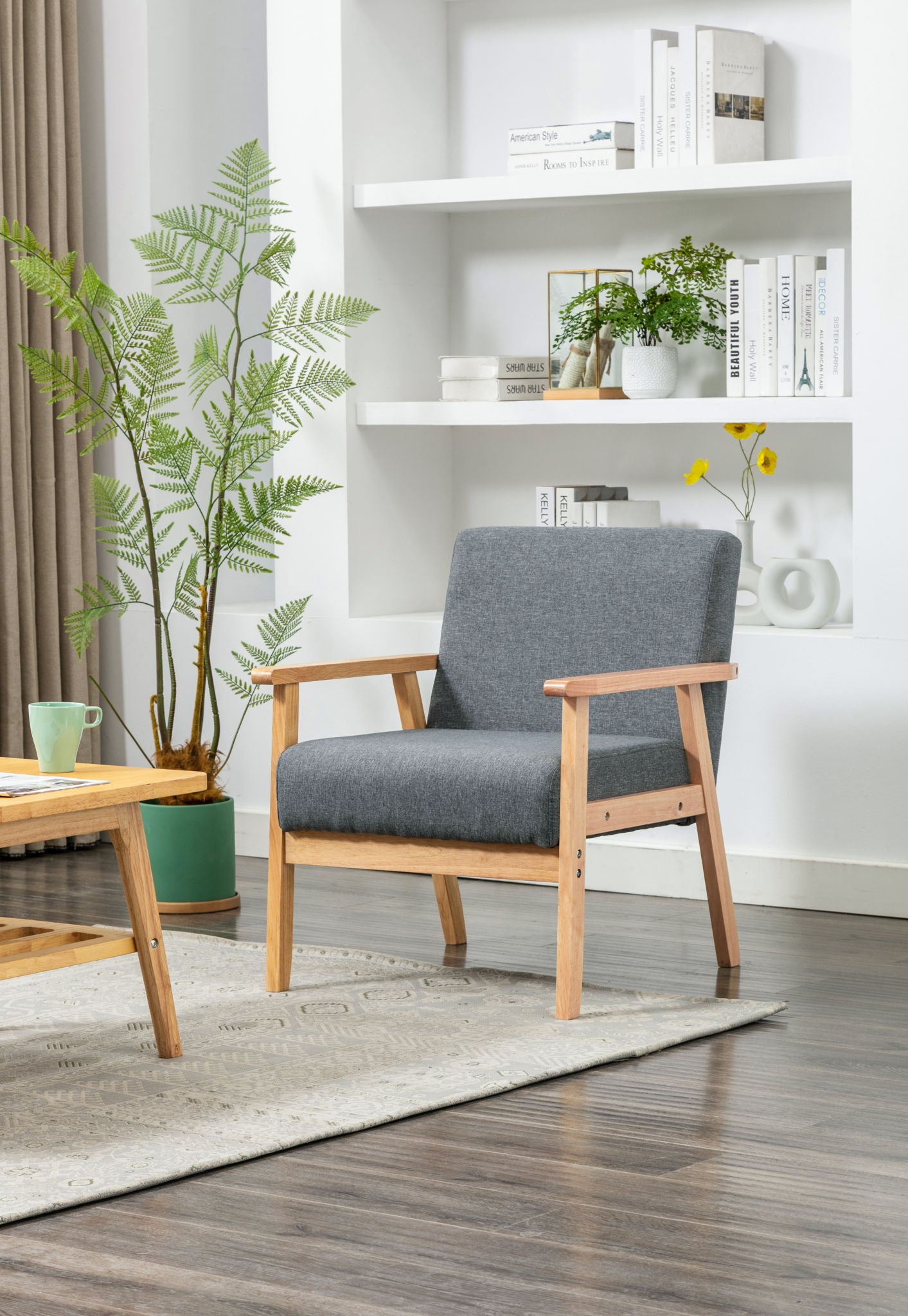 A modern living room features the Bahamas - Linen Fabric Chair in gray, a wooden coffee table with a green mug, a large potted plant, white shelves with books and decor, plus a light rug on dark wood flooring.