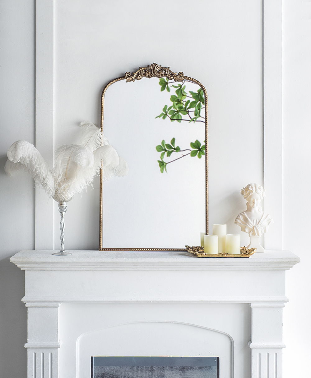 A white fireplace mantel features the Arch Mirror, Baroque Inspired Wall Decor, reflecting green leaves. The mantel is decorated with a vase of white feathers, a gold tray with candles, and a small white bust.