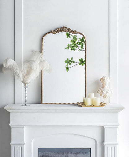A white fireplace mantel features the Arch Mirror, Baroque Inspired Wall Decor, reflecting green leaves. The mantel is decorated with a vase of white feathers, a gold tray with candles, and a small white bust.