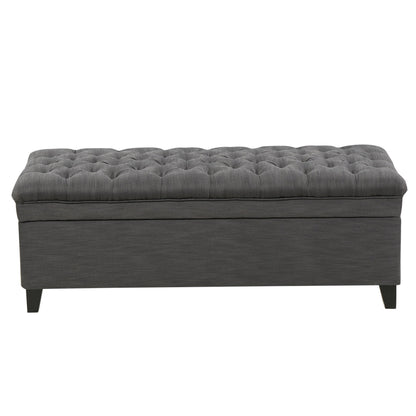 The Juliana Storage Ottoman features a rectangular shape, dark gray upholstery, tufted cushion top, and short birch legs, shown from the front.