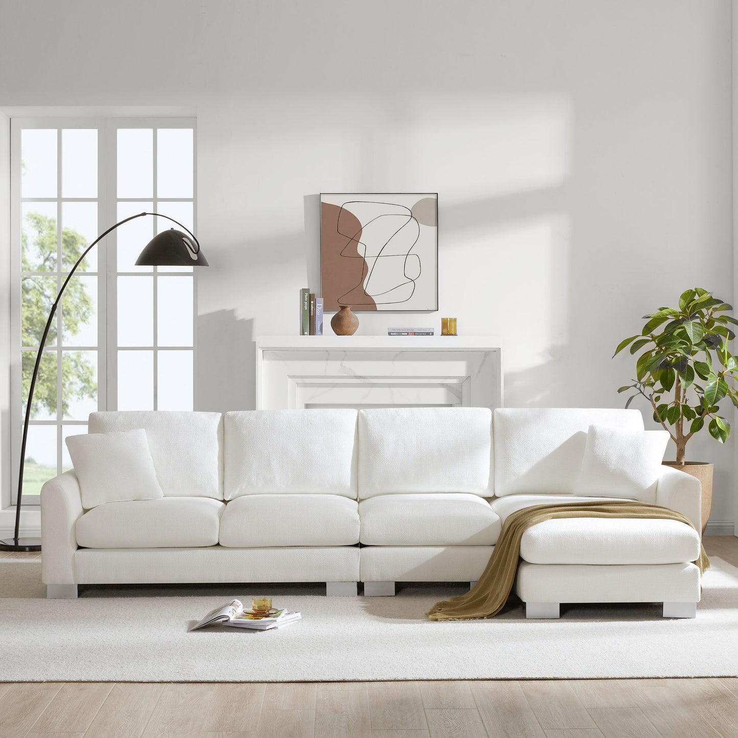 A bright living room with a Modern Oversized Sectional Sofa, two free pillows, a tan throw blanket, black floor lamp, potted plant, white fireplace, abstract wall art, and large windows letting in natural light.