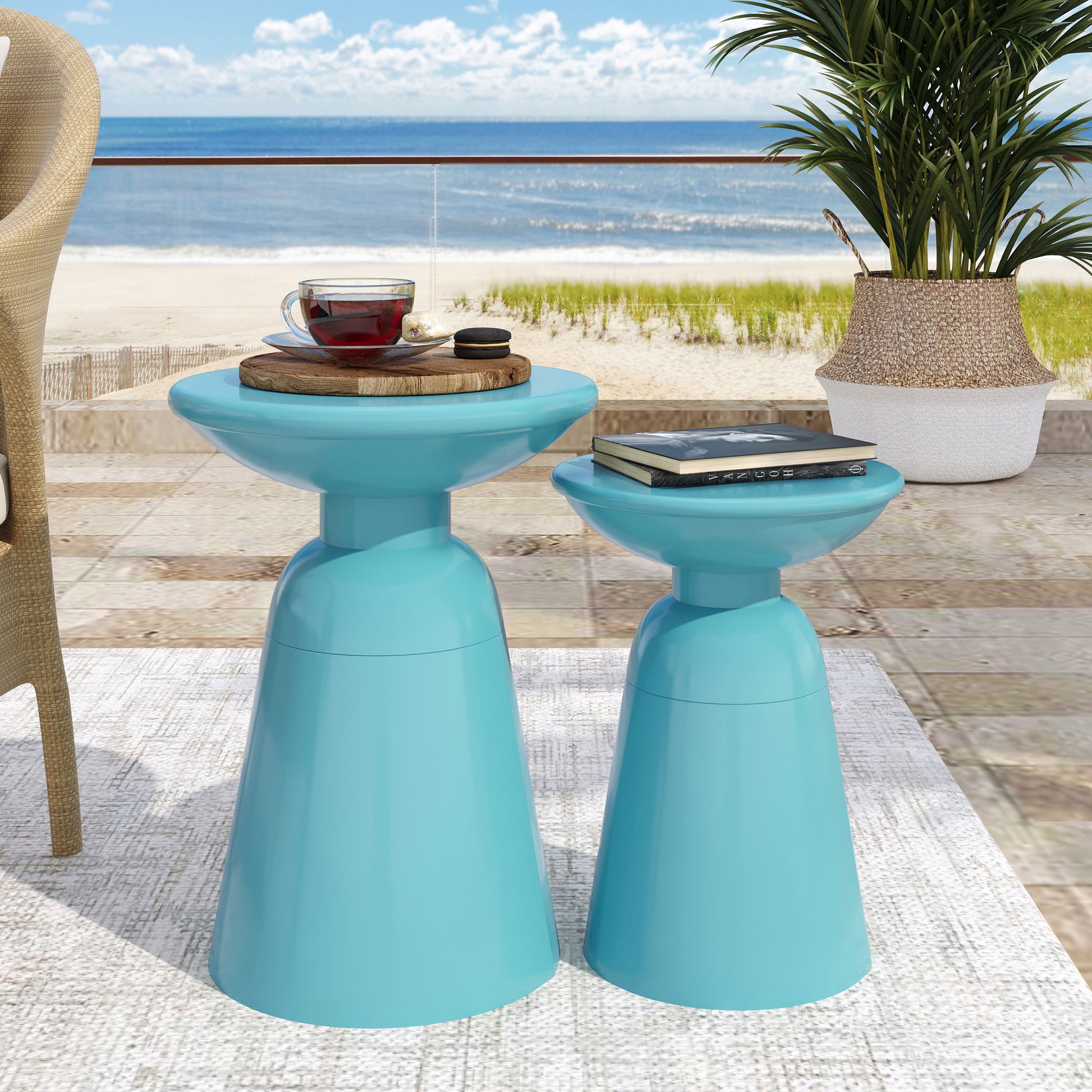 On a patio by the beach, two Serenita Outdoor Side Tables with a flared design hold a tray with teapot and cup, plus a book and sunglasses. A nearby potted plant completes the serene seaside scene.