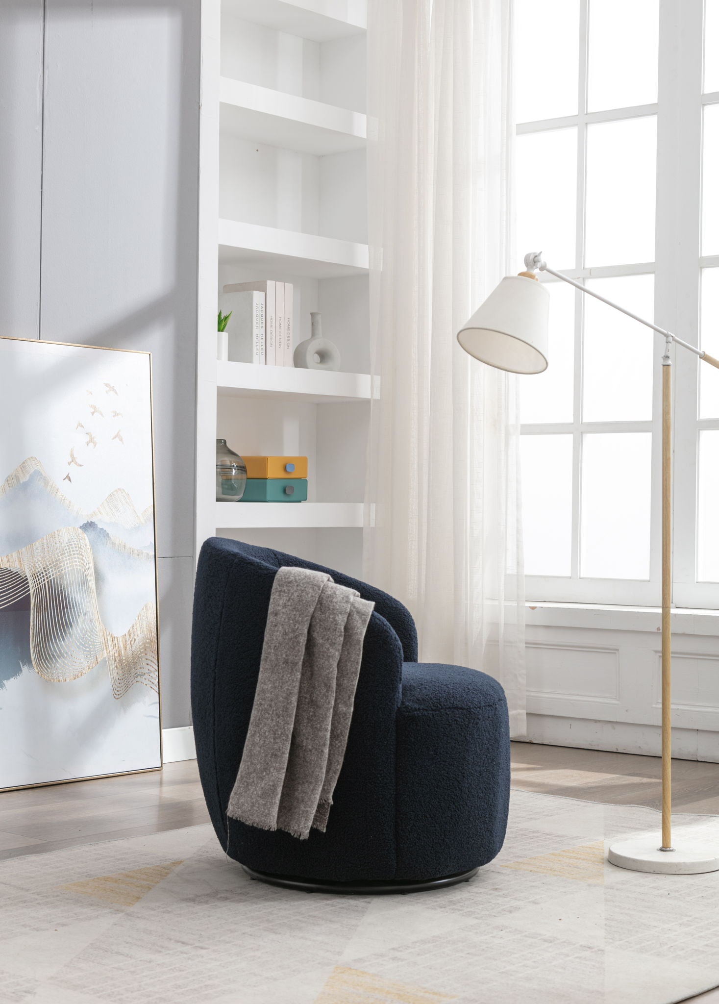 A Teddy Fabric Swivel Accent Armchair Barrel Chair with a gray blanket sits by a tall window. A white floor lamp is nearby, with minimalist shelving and books in the background, and framed abstract art resting on the floor.