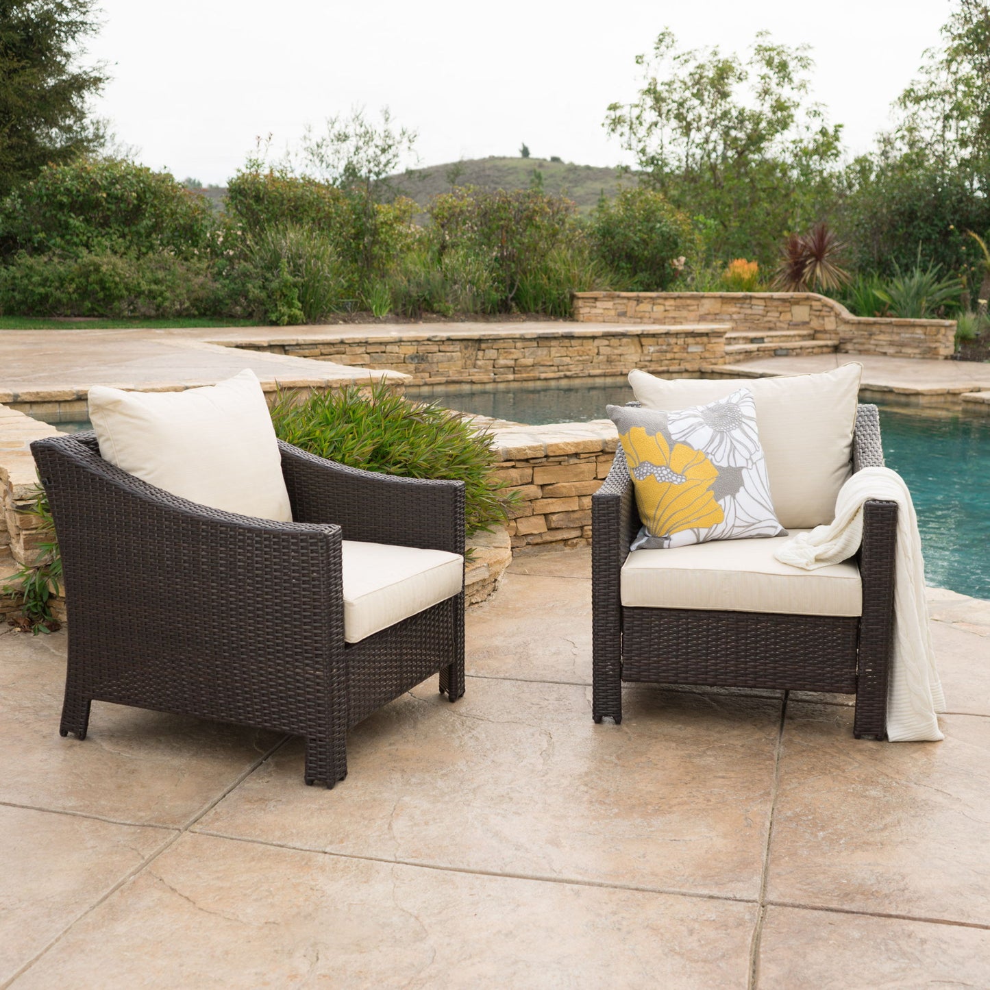 Two Antibes Armchairs with water-resistant cream cushions sit on a stone patio by a pool. One chair features a throw blanket and floral pillow. Lush greenery and a stone retaining wall complete the scene.