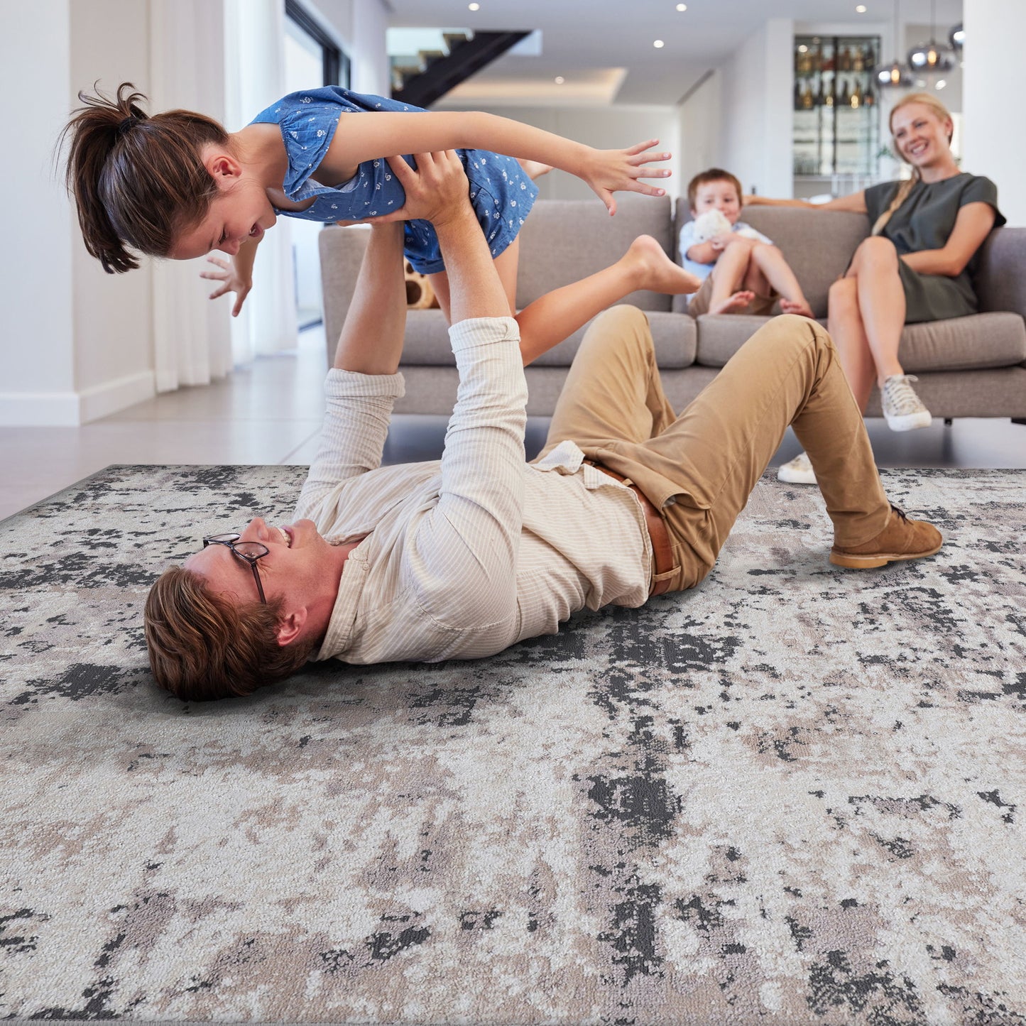 A man lies on the Payas - Abstract Area Rug, lifting a smiling young girl in a blue dress. In the background, a woman and boy watch from a gray sofa. The scene is set in a bright, modern living room.