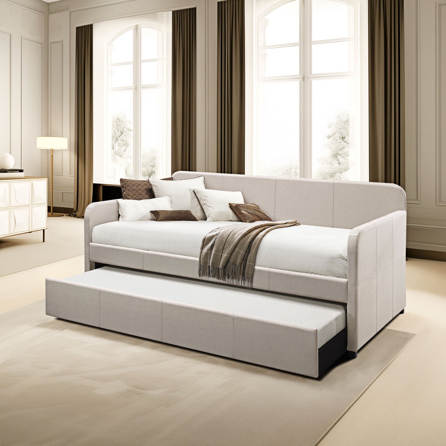 A modern living room features the Jagger - Twin Daybed With Trundle in Fog, adorned with pillows and a brown-striped throw, set against large windows with long curtains. The daybed includes a convenient pull-out trundle bed.