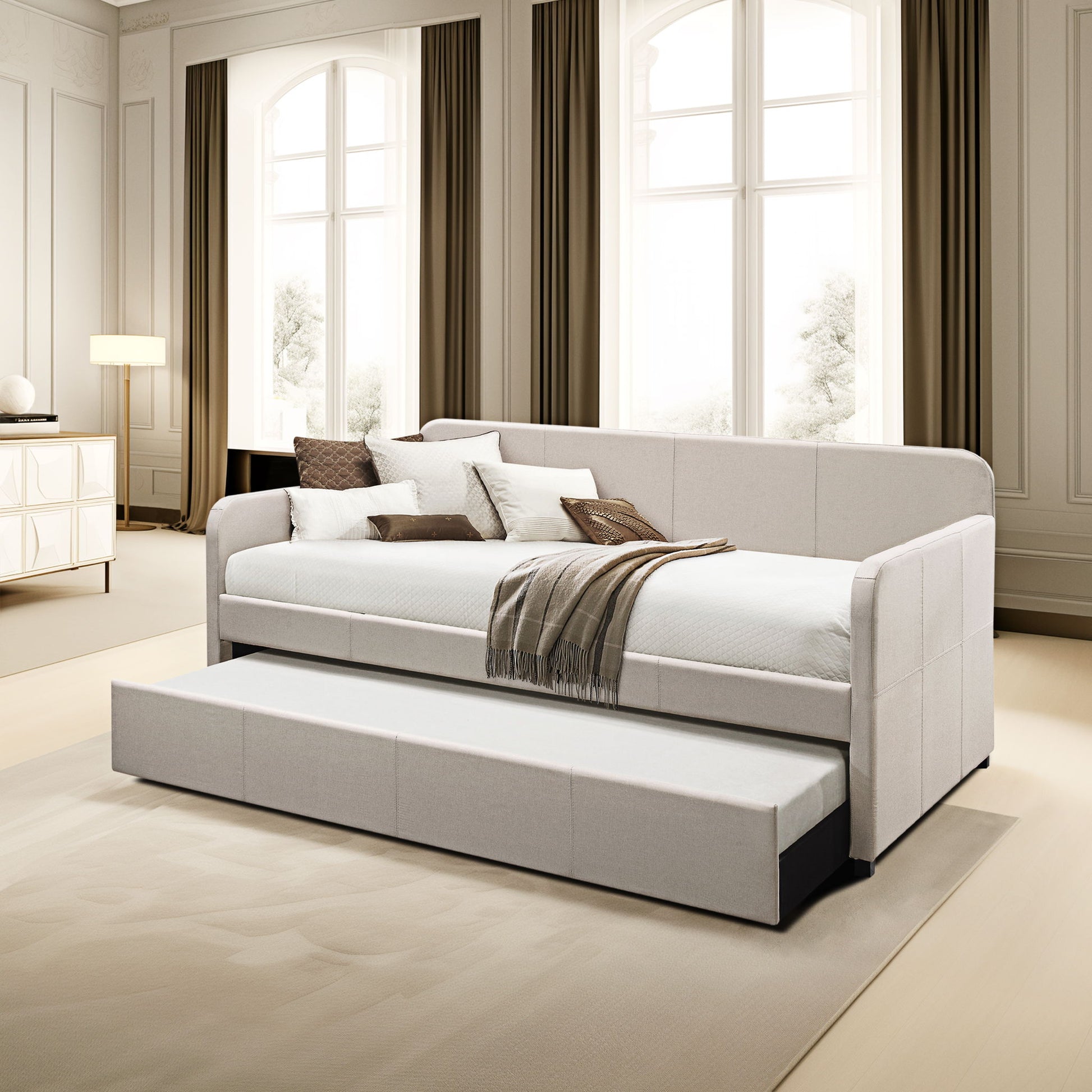 A modern living room features the Jagger - Twin Daybed With Trundle in Fog, adorned with pillows and a brown-striped throw, set against large windows with long curtains. The daybed includes a convenient pull-out trundle bed.