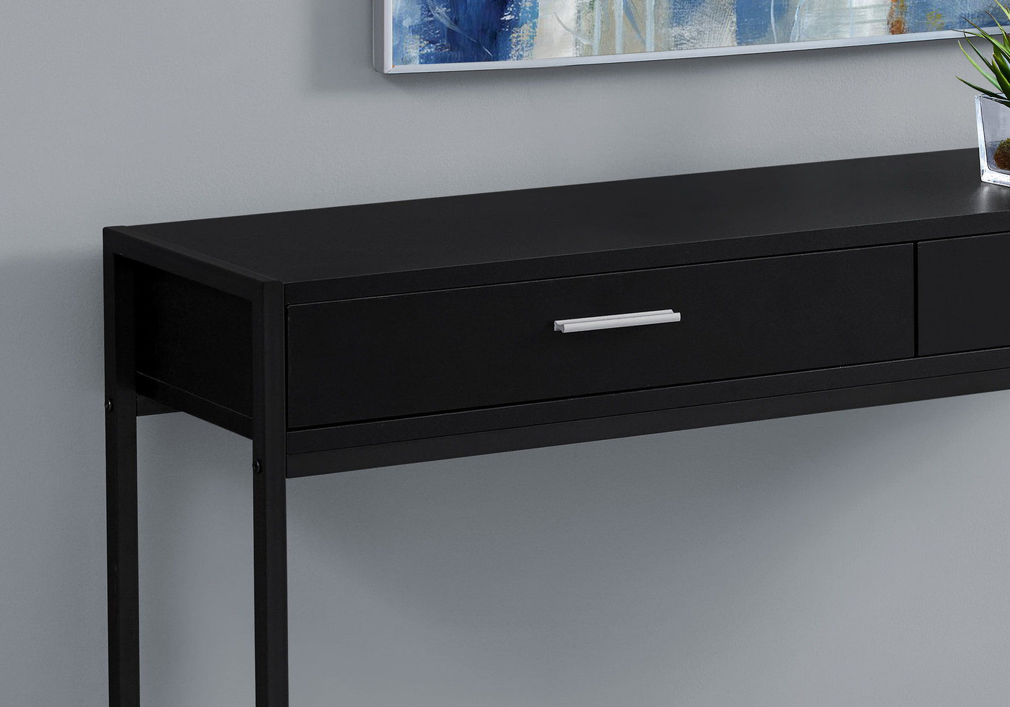 The Accent Console Table For Entryway, featuring storage drawers in a contemporary black design with a silver handle, is set against a light gray wall. A framed artwork and a small potted plant sit on top.