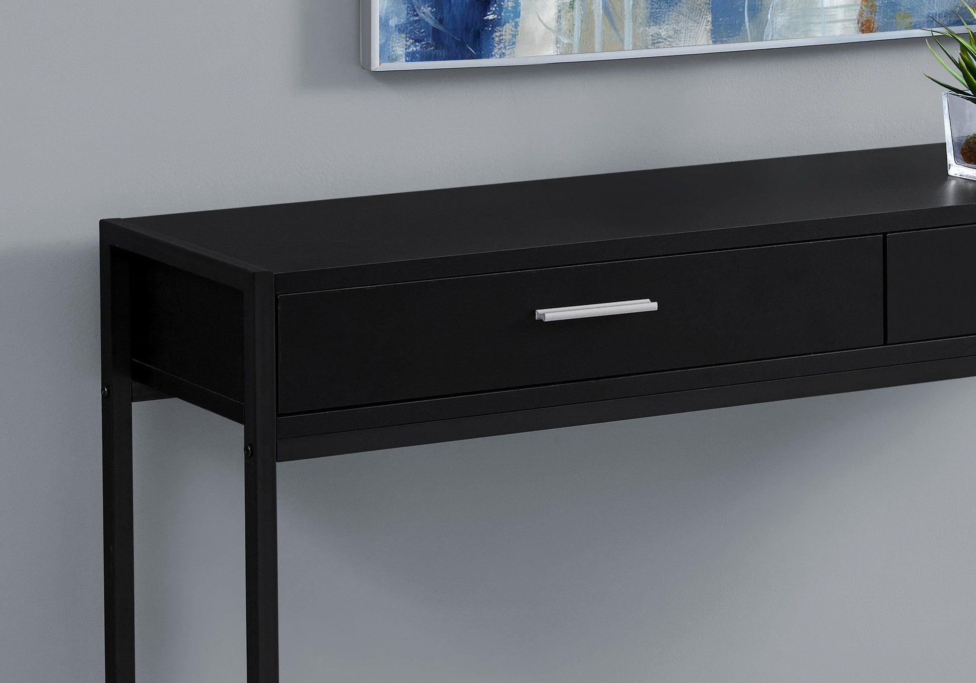 The Accent Console Table For Entryway, featuring storage drawers in a contemporary black design with a silver handle, is set against a light gray wall. A framed artwork and a small potted plant sit on top.