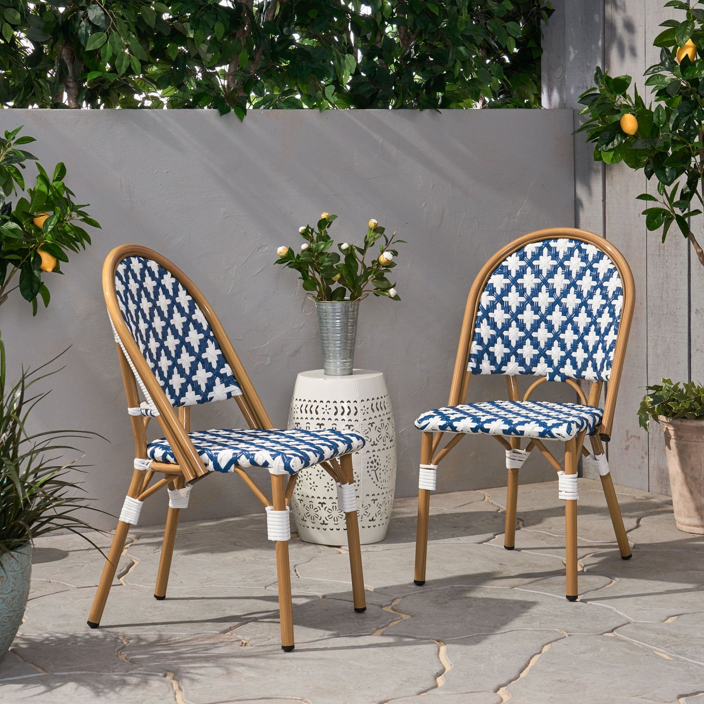 Two Chic French Bistro Chairs with durable rattan and aluminum frames, featuring blue and white patterns, sit on a stone patio among green plants, potted lemons, and a small white side table topped with a vase of white flowers.