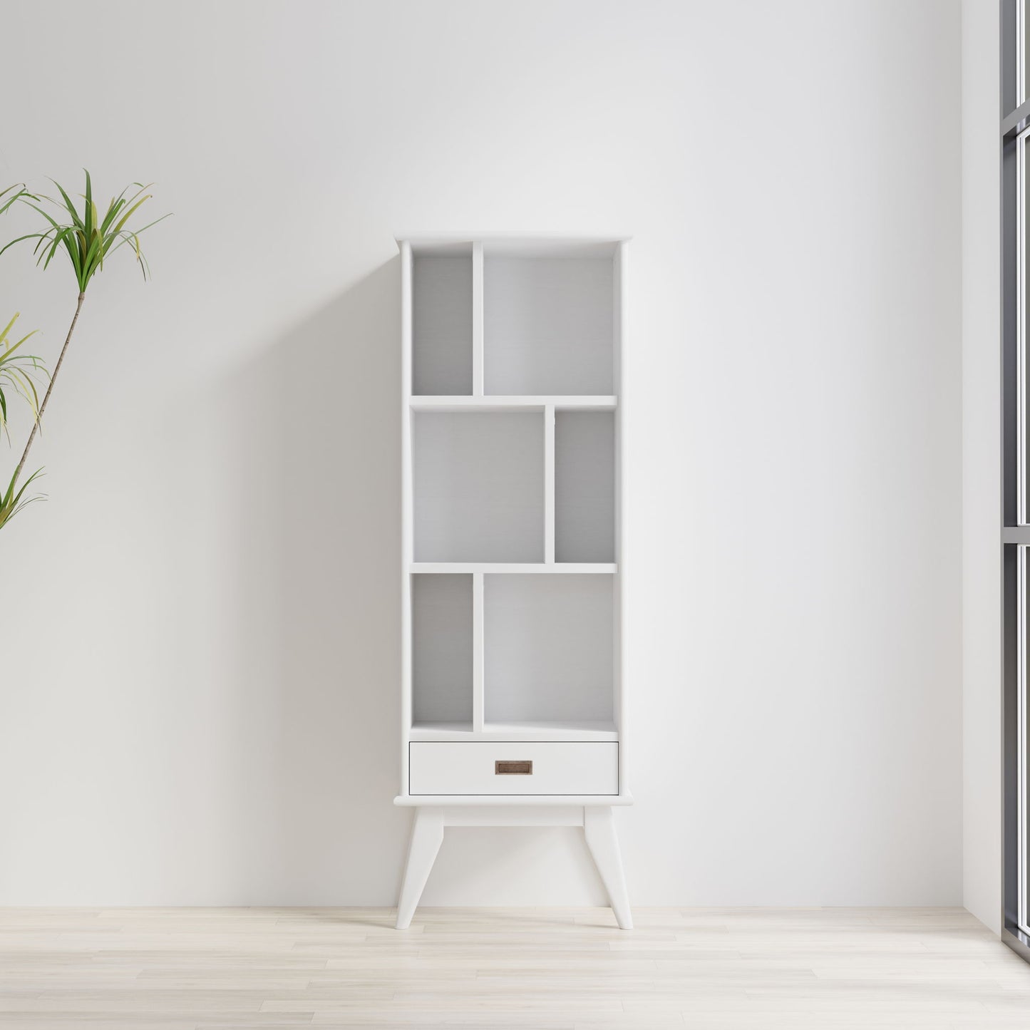 The Draper - Mid Century Wide Bookcase And Storage Unit, featuring geometric shelves and a single bottom drawer, stands empty against a white wall in a bright minimalist room with light wood floors and a tall potted plant to the left.
