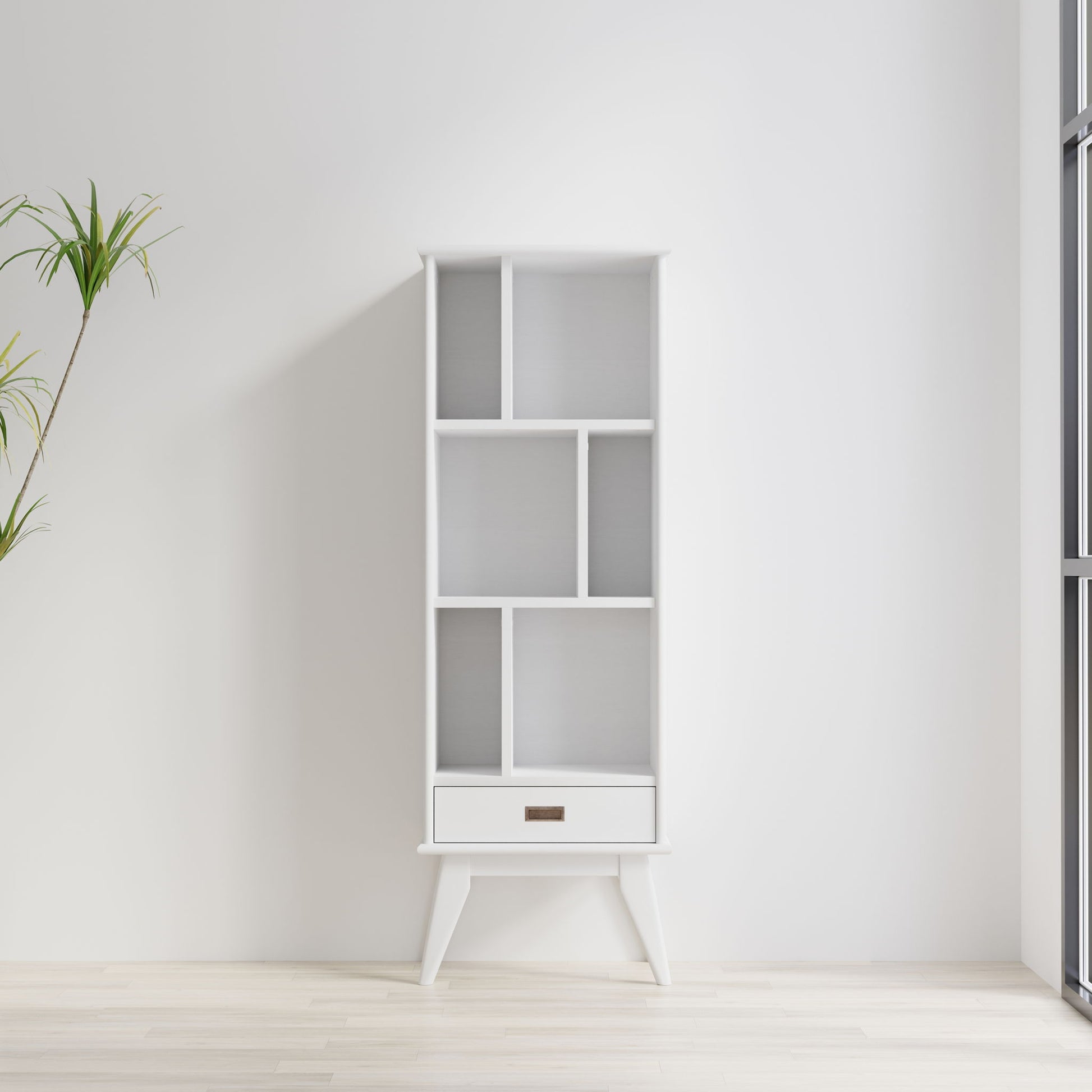 The Draper - Mid Century Wide Bookcase And Storage Unit, featuring geometric shelves and a single bottom drawer, stands empty against a white wall in a bright minimalist room with light wood floors and a tall potted plant to the left.