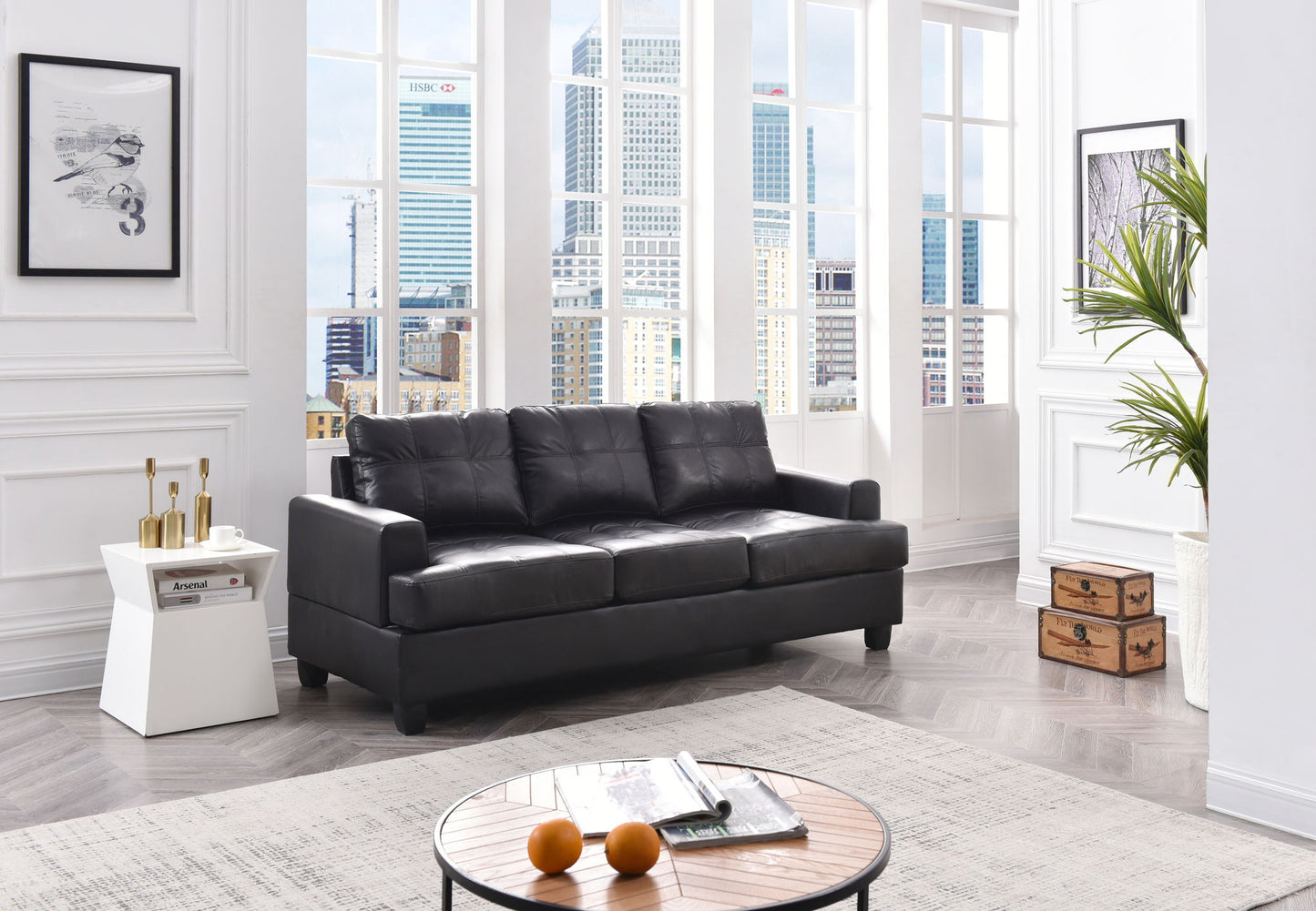A modern living room featuring the Sandridge sofa in black leather, decorated with stylish pillows. A white-gold side table sits beside potted plants and framed art, while oranges rest on a round coffee table by large windows revealing city views.