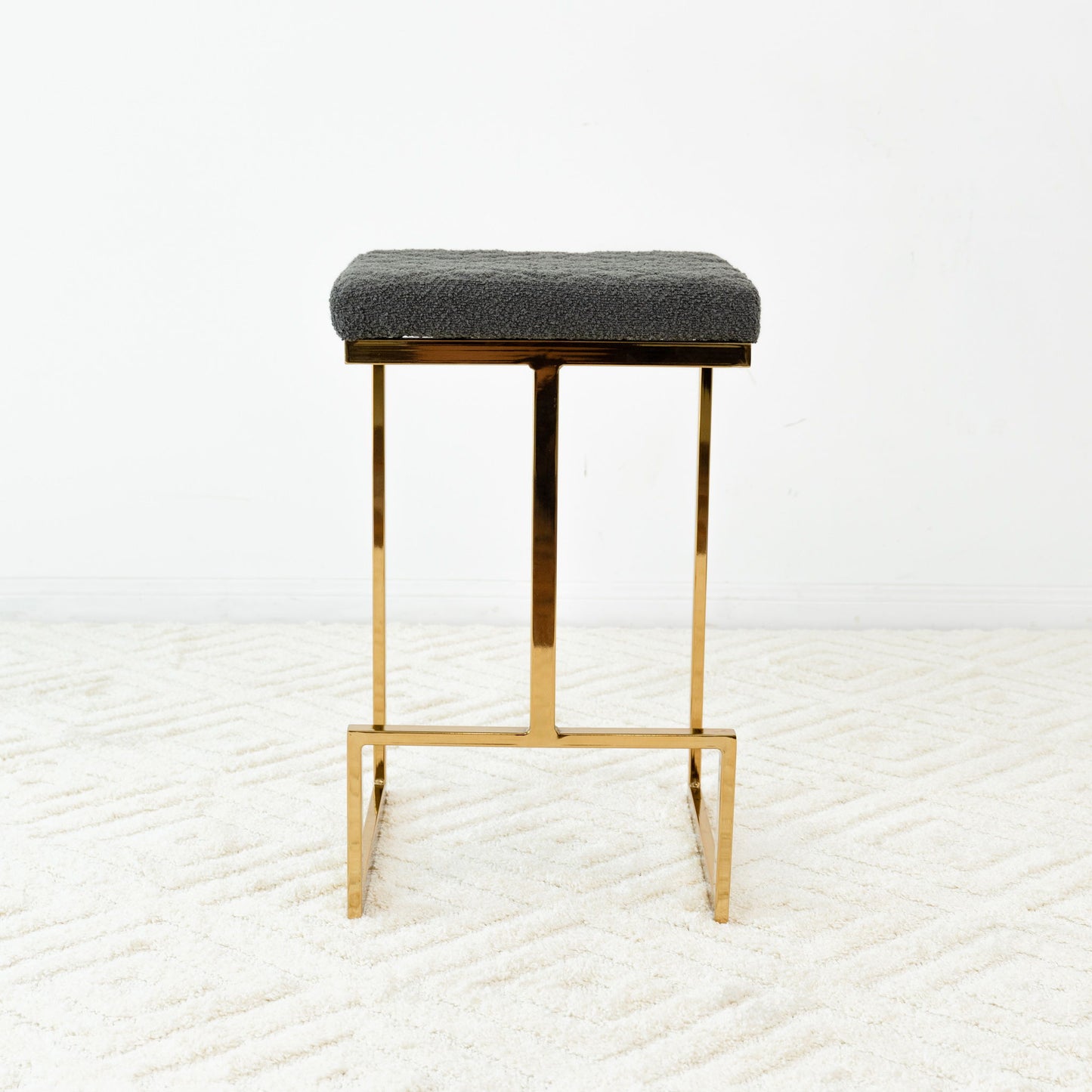 The Joel - Mid Century Modern Luxury Upholstered Stool, featuring a gold metal frame and rectangular textured dark gray cushion, sits on a white patterned carpet against a plain white wall.