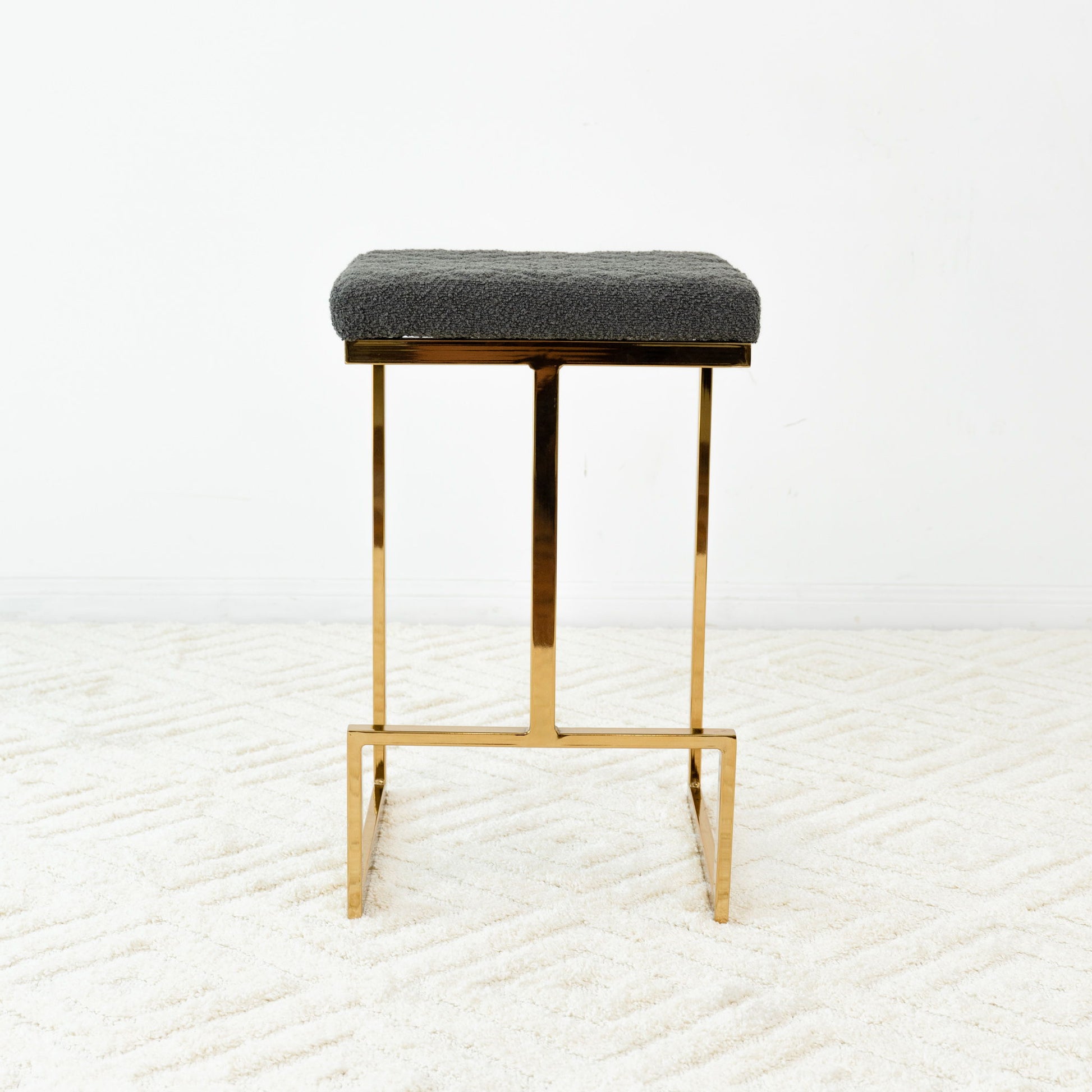 The Joel - Mid Century Modern Luxury Upholstered Stool, featuring a gold metal frame and rectangular textured dark gray cushion, sits on a white patterned carpet against a plain white wall.