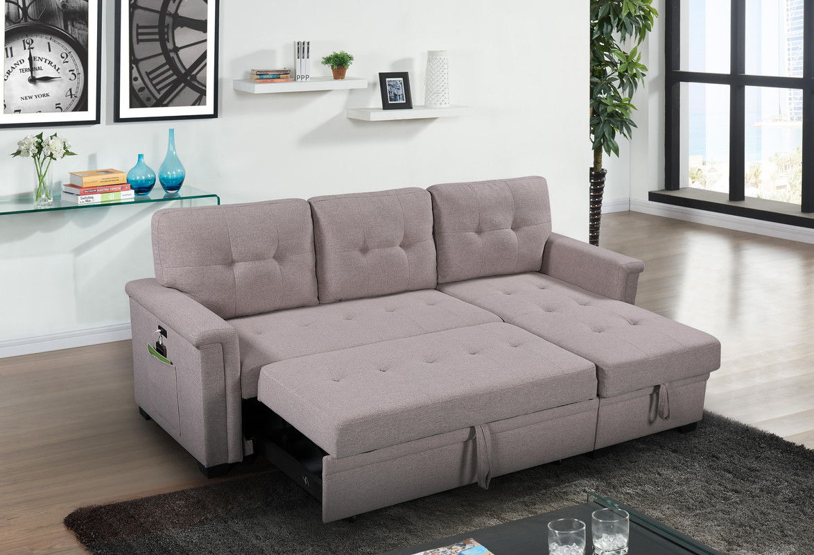 The Ashlyn Reversible Sleeper Sectional Sofa with storage chaise, USB charging ports, and pocket is unfolded in a modern living room with wooden floors, a glass coffee table, wall shelves, vases, and a large clock artwork.