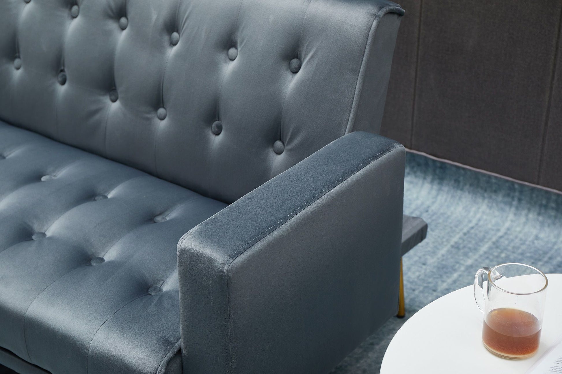 A close-up of the Comfort Compact Design Sofa Bed in dark blue, placed beside a small white round table holding a clear glass mug with tea or coffee on a blue textured carpet.