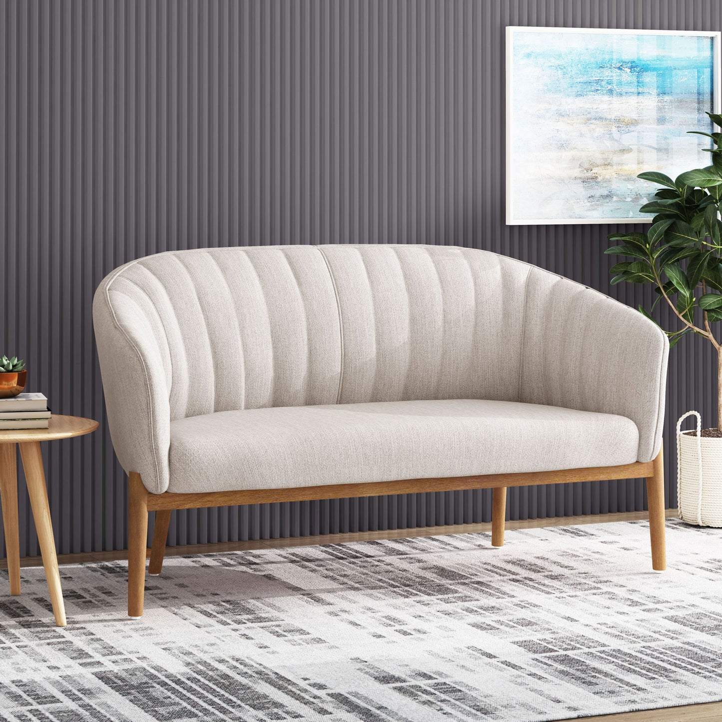 The Mid Century Modern Loveseat With Channel Stitched Backrest in beige sits on a patterned rug; to the left is a round wooden side table with books, to the right is a potted plant, and a framed abstract artwork hangs on the gray textured wall behind.