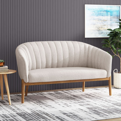 The Mid Century Modern Loveseat With Channel Stitched Backrest in beige sits on a patterned rug; to the left is a round wooden side table with books, to the right is a potted plant, and a framed abstract artwork hangs on the gray textured wall behind.