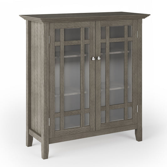 The Bedford - Storage Media Cabinet features a gray wooden finish, glass-paneled double doors with silver knobs, two interior shelves, and stands on four legs against a plain white background.