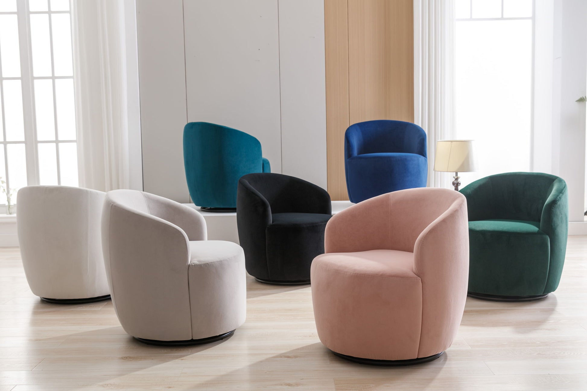 Six Velvet Fabric Swivel Accent Armchair Barrel Chairs with powder-coated metal rings in white, pink, green, black, teal, and blue are arranged in a bright room with large windows, light wood floors, and neutral walls.