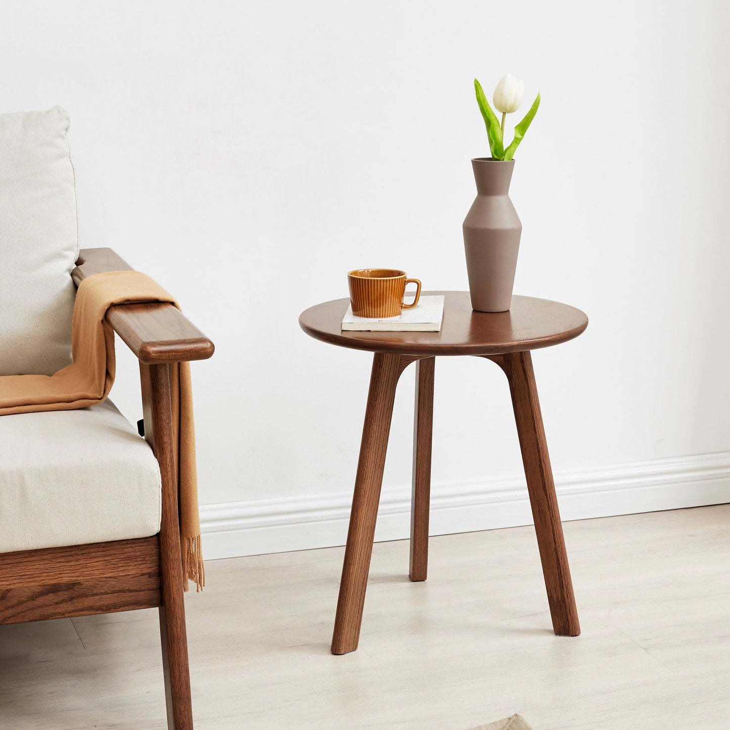 A Round Small End Table, Side Table For Bedroom & Balcony with three legs holds a brown cup, saucer, and a beige vase with a white tulip. Beside it is a wooden sofa featuring a beige cushion and a light brown throw on the armrest.