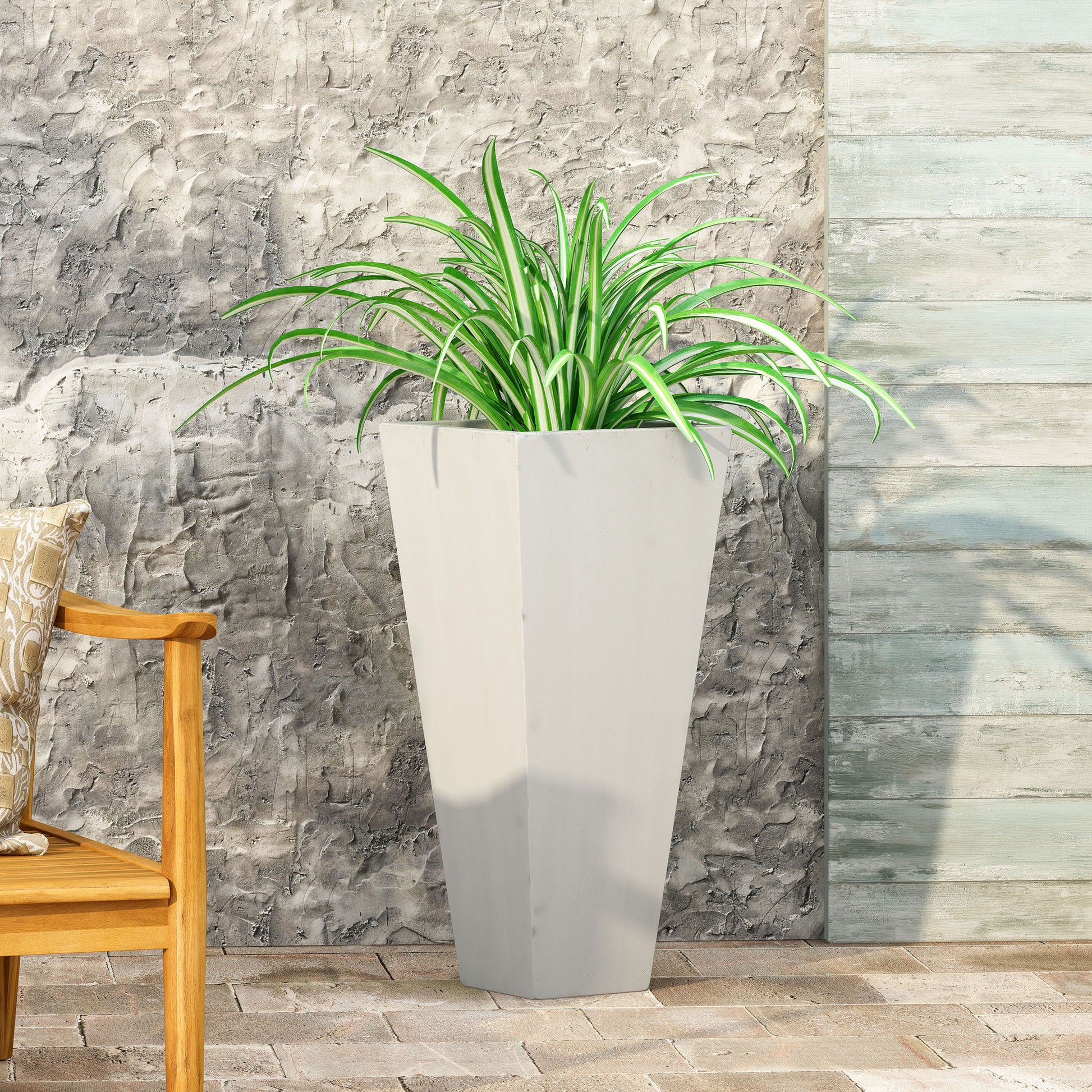 An Outdoor Modern Cast Stone Planter in light gray with a spider plant sits on a stone patio by a textured gray wall. On the left, part of a wooden bench with a patterned pillow is visible.