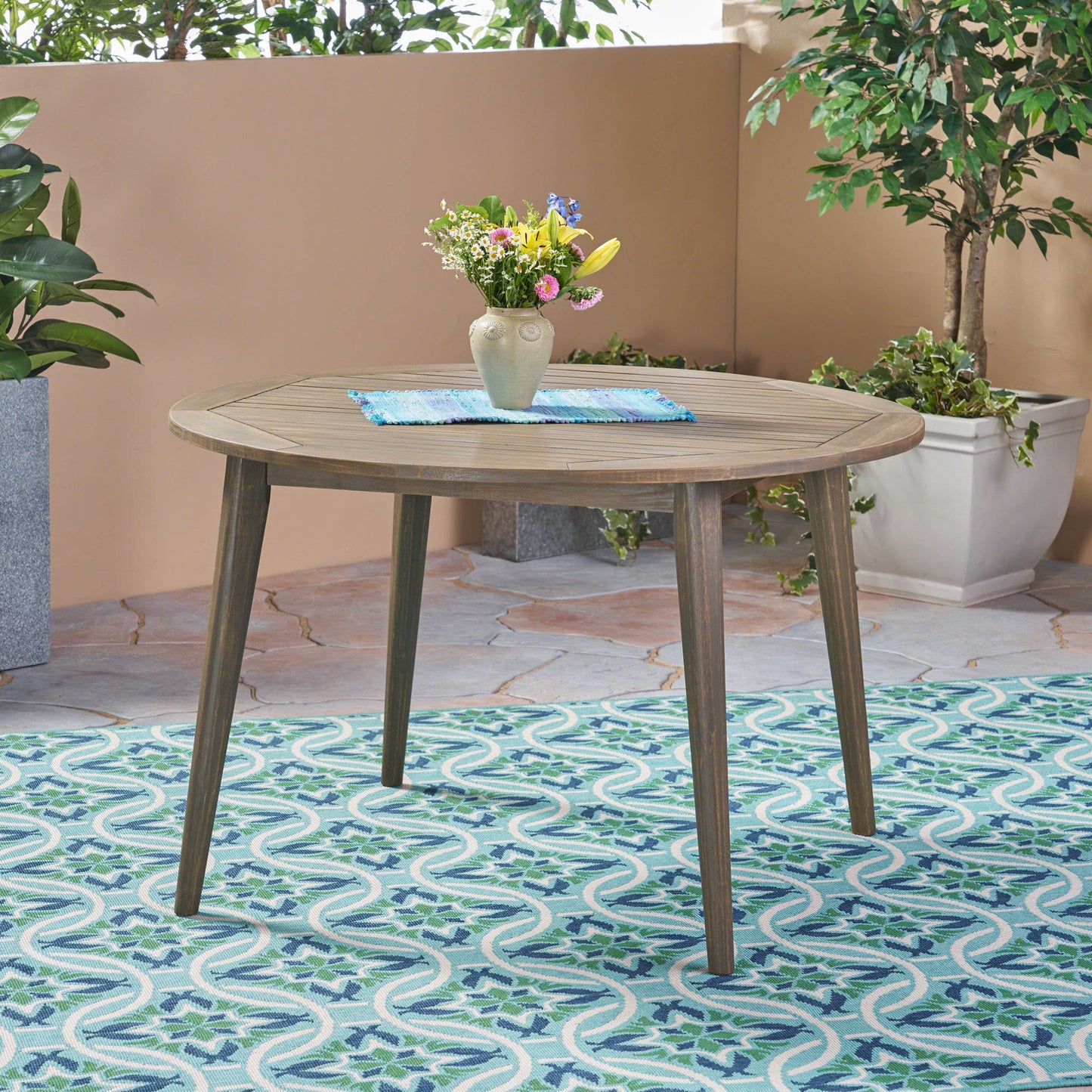 The Stamford Outdoor Dining Table, featuring a modern design, is paired with vibrant flowers and potted plants on a patterned blue-green rug in an inviting patio setting.