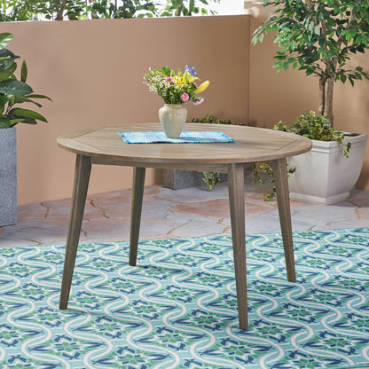 The Stamford Outdoor Dining Table, featuring a modern design, is paired with vibrant flowers and potted plants on a patterned blue-green rug in an inviting patio setting.