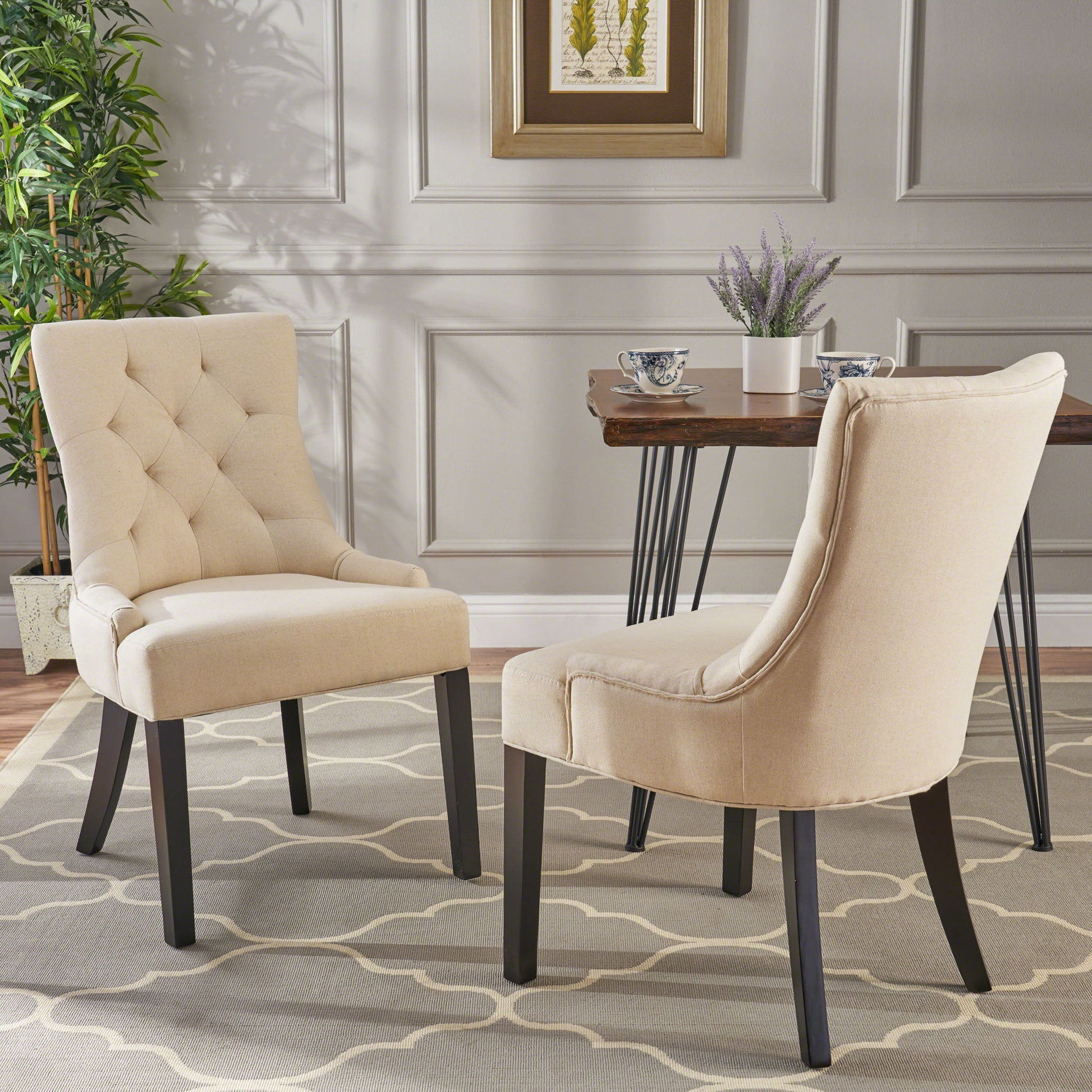 A set of Cheney Contemporary Dining Chairs with tufted beige upholstery and dark wooden legs sits on a patterned rug in a stylish room, featuring a table with teacups, a potted plant, paneled walls, and framed artwork.