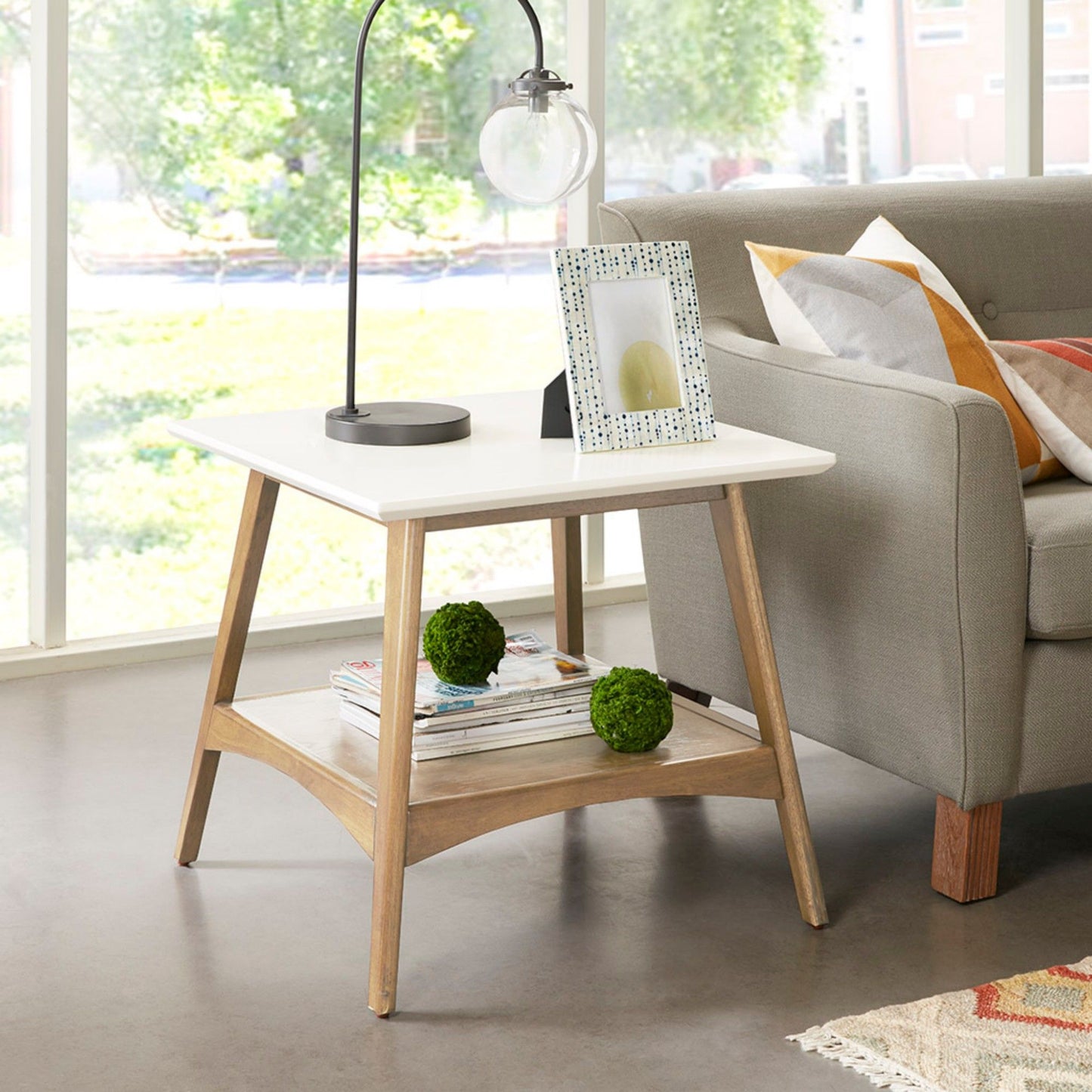 The Modern Table With Extra Storage with a white top and wooden legs sits by a gray sofa. A framed photo, lamp, and books are on it, with two green moss balls below. Large windows fill the background with light.