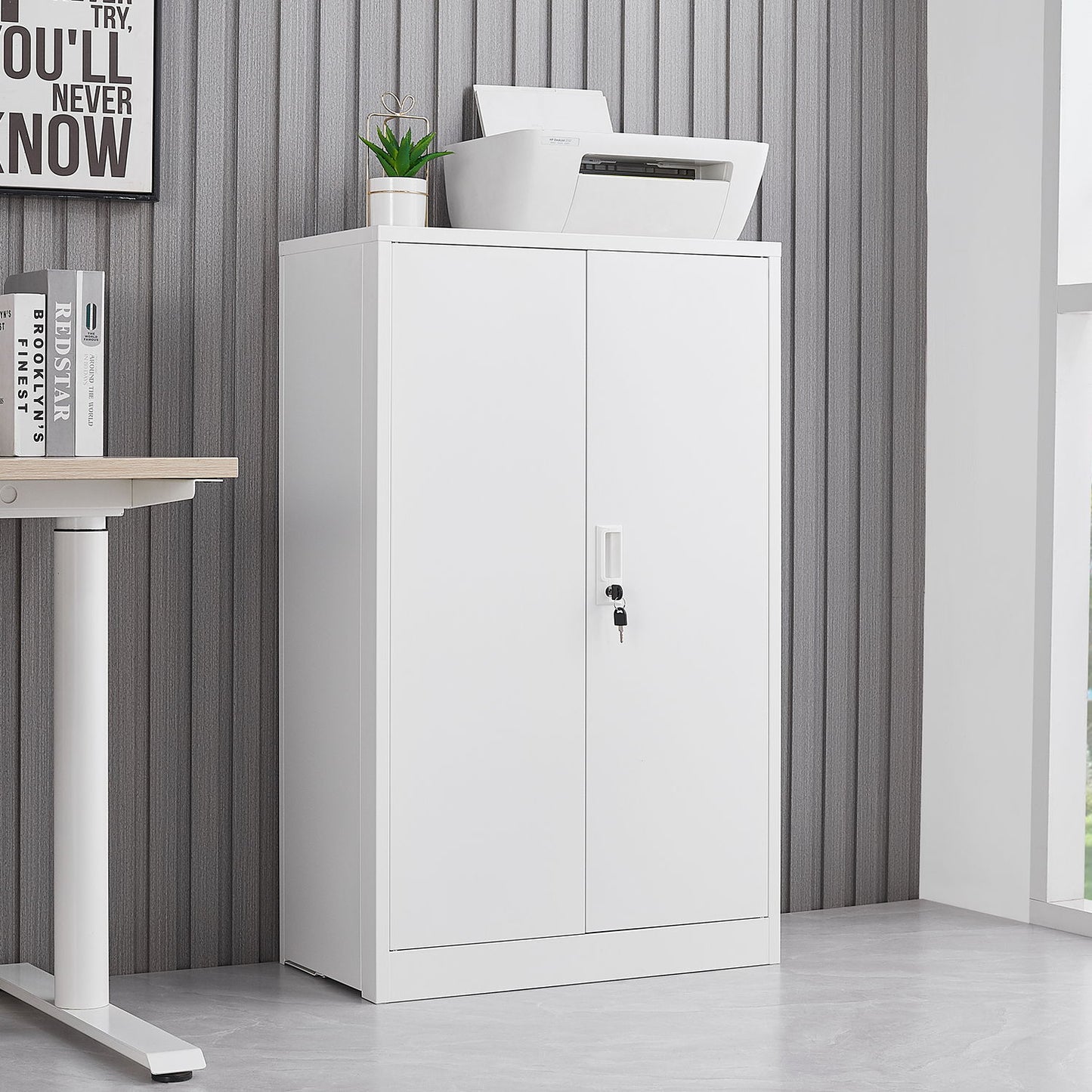 A Metal Storage Cabinet With Locking Doors And Adjustable Shelf stands against a gray striped wall. A printer, potted plant, and framed sign sit on top, next to a white desk on the left. This folding storage locker is ideal for home offices or garages.