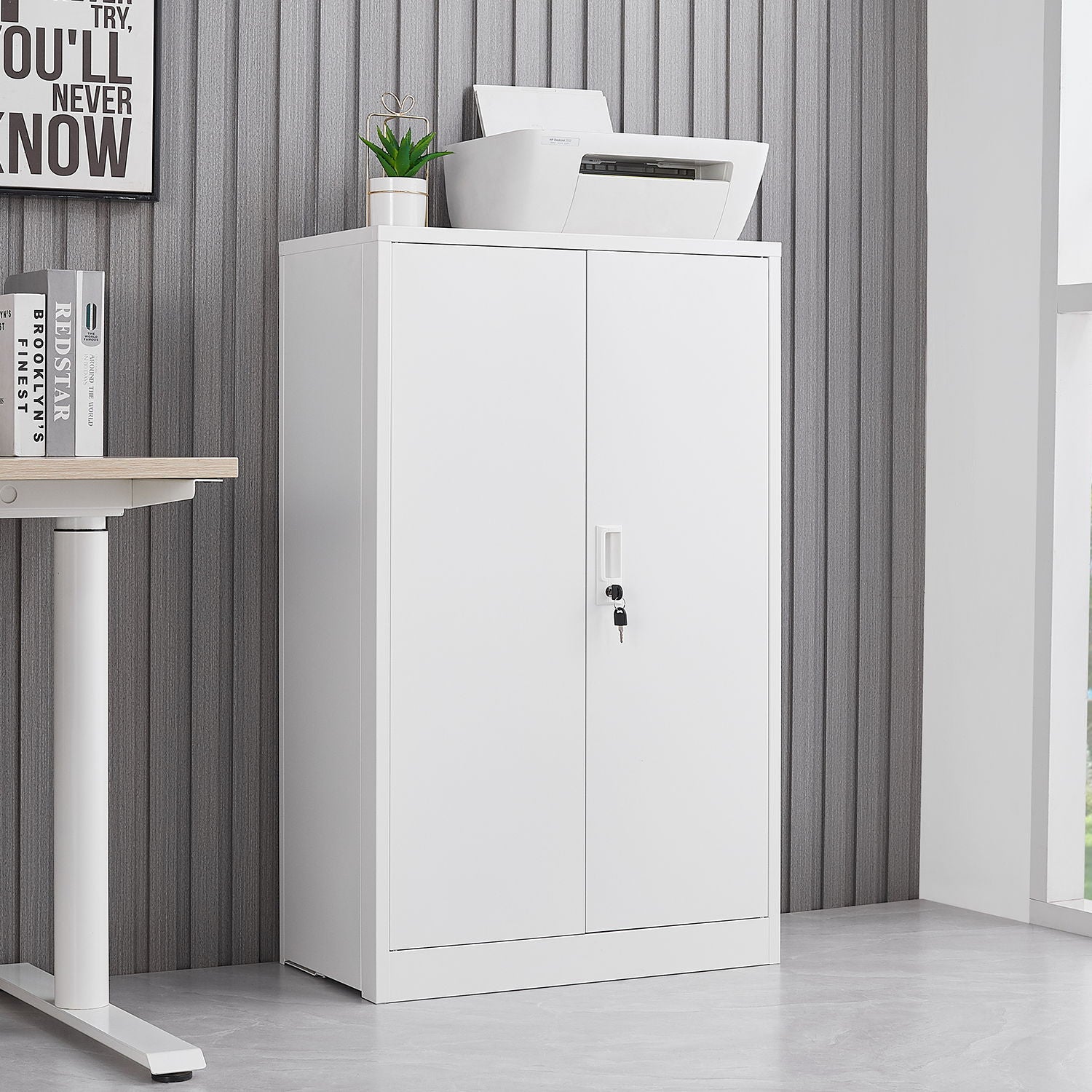 A Metal Storage Cabinet With Locking Doors And Adjustable Shelf stands against a gray striped wall. A printer, potted plant, and framed sign sit on top, next to a white desk on the left. This folding storage locker is ideal for home offices or garages.