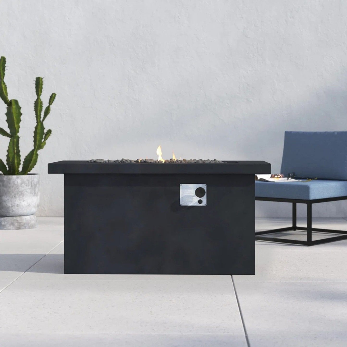 A sleek Propane / Gas Outdoor Fire Pit Table with a small flame sits on a light patio, near a potted cactus and a blue cushioned bench.